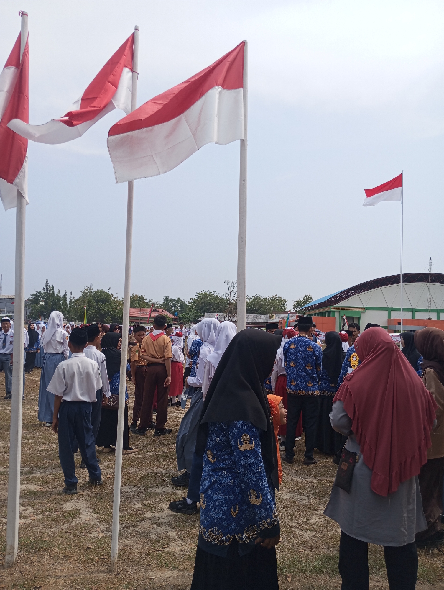 The image depicts an outdoor gathering of people, primarily students, in what appears to be a field or open ground. Three Indonesian flags, red and white, are prominently displayed on tall poles in the foreground. One flag is closer to the camera and appears to be partially unfurled, while two others are further away and more visible. A smaller Indonesian flag is visible in the background, mounted on a pole near a building.
The crowd consists of individuals of various ages, many of whom are wearing school uniforms. The uniforms include white shirts and dark trousers or skirts, with some students also wearing headscarves. Some individuals are wearing scout uniforms. The general atmosphere suggests an organized event, possibly a flag ceremony or a school assembly.
The environment is characterized by dry, grassy ground with scattered patches of dirt. In the background, there is a large building with a distinctive arched roof, which appears to be a sports facility or a public building. Trees and other vegetation are visible in the distance, as well as some smaller structures. The sky is overcast. The overall scene suggests a location in Pematang Reba, Indonesia, likely a school or a public area designated for events.