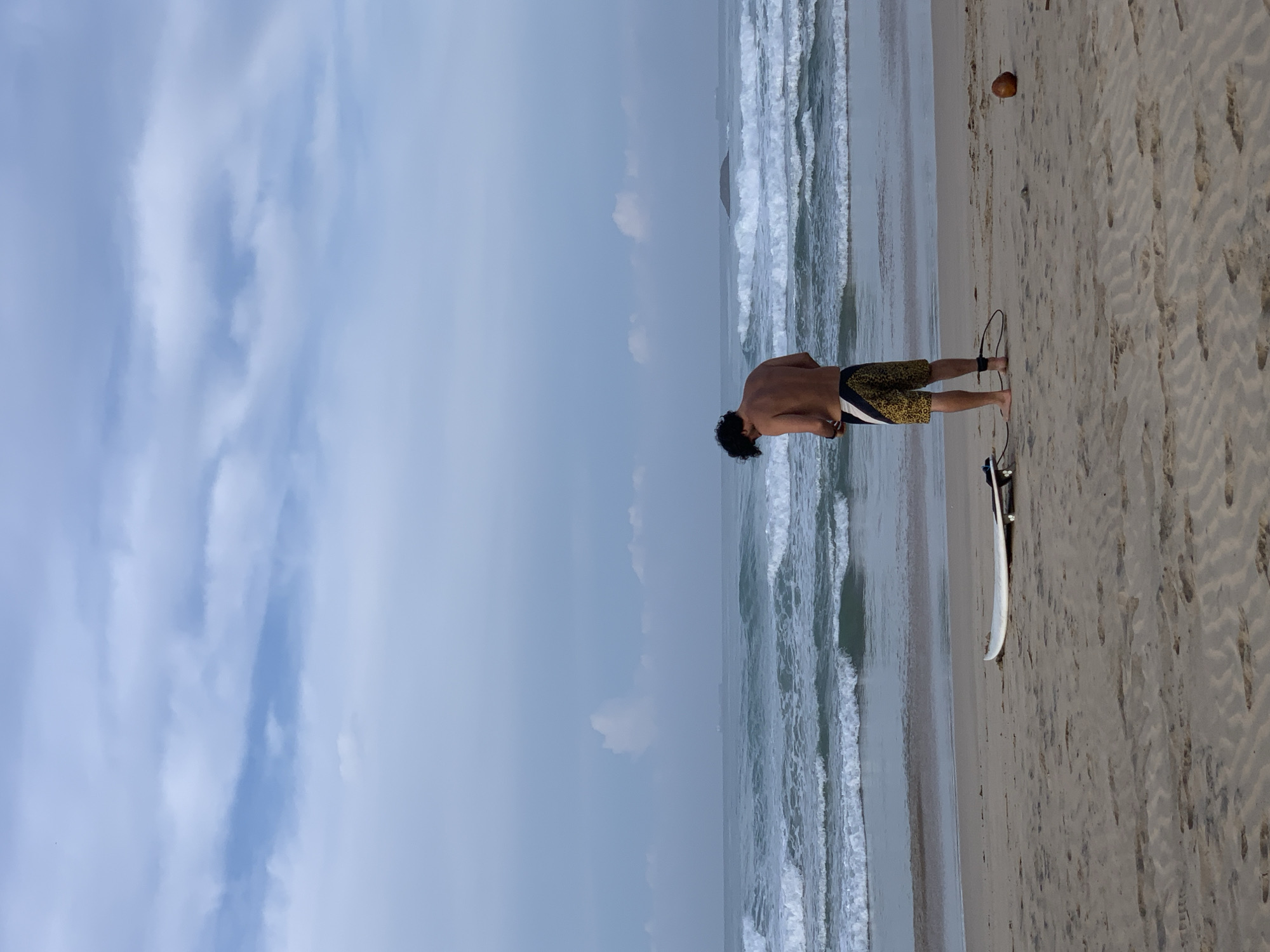 A man stands on a beach in Kathaluwa, Sri Lanka, with his back to the camera, facing the ocean. He has short, dark hair and wears patterned swim trunks with a white stripe on the side. Next to him, on the sand, lies a surfboard with a leash attached. Further up the beach, a coconut rests on the sand. The ocean stretches out in front of him with visible waves. Above, the sky is a mix of light blue and white with a cloudy overlay.