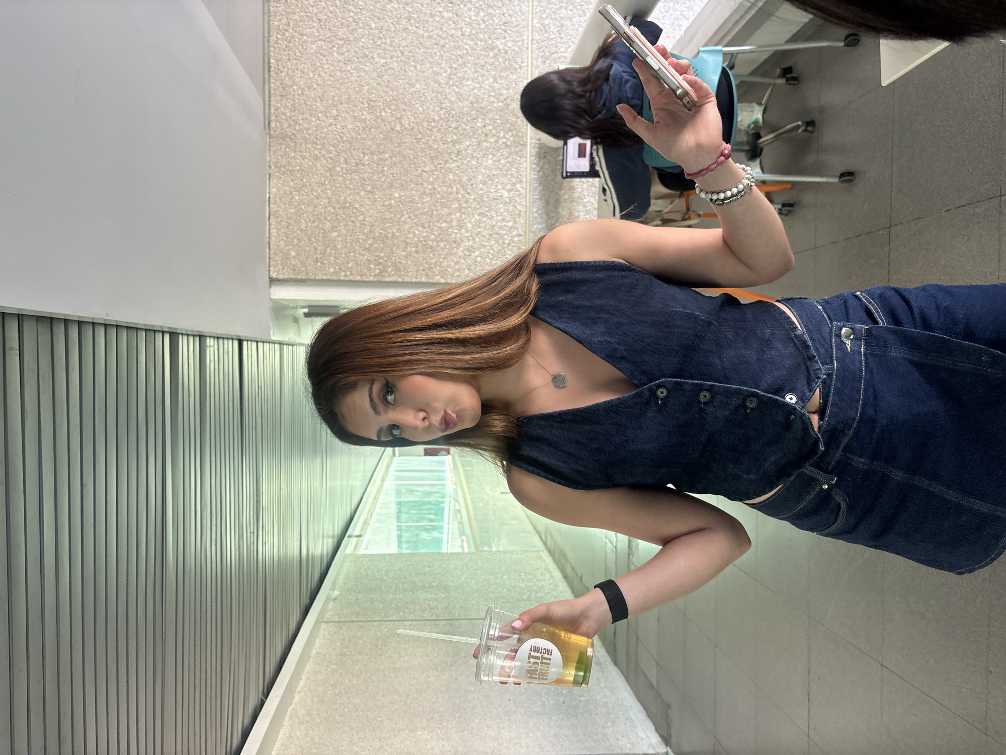 The image shows a young woman in Puebla, Mexico, posing for a photograph in a hallway. She is wearing a dark denim outfit consisting of a vest and skirt or shorts. She holds a clear plastic cup with a straw in her left hand, and her right hand is raised, holding a cell phone as if taking a selfie. She has light brown hair with blonde highlights and is wearing a necklace and bracelets. Behind her, another person is seated at a desk with a laptop, and other people are in the background. The environment appears to be an interior hallway with light-colored walls and some greenery visible outside a window.