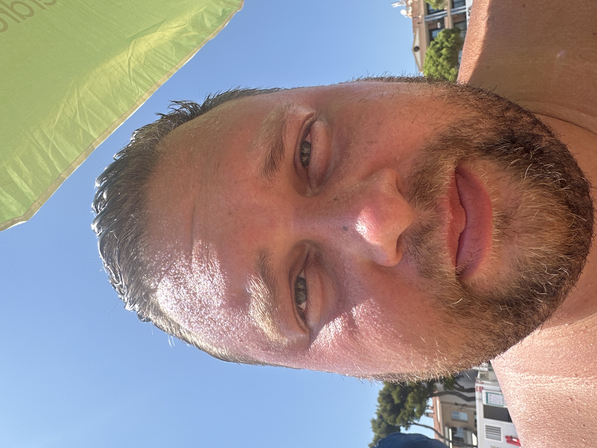 A close-up, selfie-style photograph features a man's face from the nose up, taken outdoors on a sunny day. The man has fair skin, a light brown beard, and dark, wet hair. He appears to be sweating, as evidenced by the sheen on his forehead and cheeks. A portion of a green umbrella is visible at the top right of the frame, casting a shadow over part of his face. In the background, out of focus, are buildings and trees, suggesting an urban or semi-urban environment. The sky above is a clear, bright blue. The overall impression is of a person at a beach or outdoor recreational area, likely in Sant Feliu de Guíxols, Spain, enjoying warm weather.