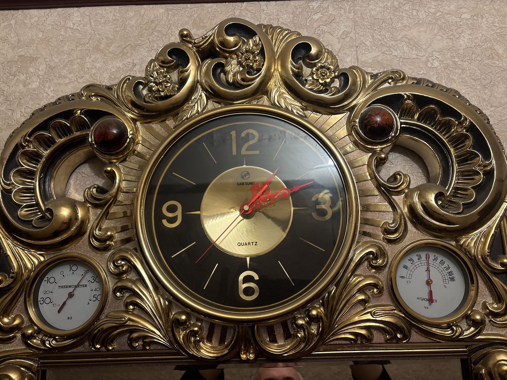 A close-up shot captures a decorative Samsung quartz clock mounted against a beige-patterned wall. The clock, encased in an ornate gold-colored frame, features a black face with gold numerals. The hands of the clock are red. A thermometer and hygrometer are set on either side of the clock face, built into the gold frame. The frame has intricate designs with floral patterns and brown jewel-like embellishments. The reflection of a person, likely the photographer, can be seen in the mirror at the bottom of the clock. There is no indication of location in Krasnoyarsk, Russia.