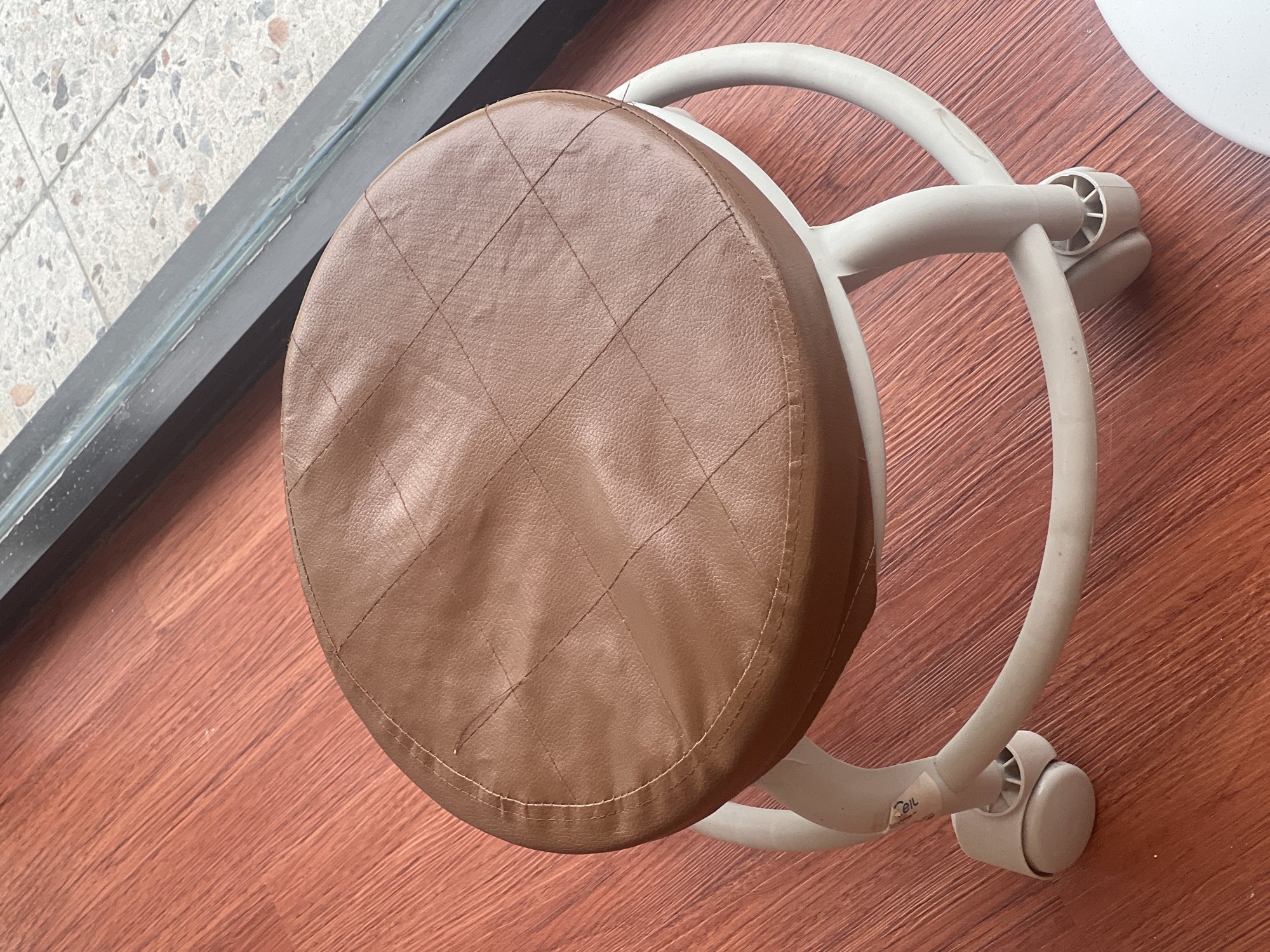 This is an overhead shot of a stool with a brown, quilted, leatherette seat and a light gray plastic base with wheels. The stool is positioned on a wooden floor with a reddish-brown hue. To the left of the stool, there's a dark gray metallic frame, likely part of a window or door, with a tiled floor visible beyond it. The tiles are a mix of white, gray, and speckled with dark flecks. The lighting suggests it is daytime, and the setting appears to be indoors, possibly in a home or commercial space in Gianyar, Indonesia. There is no discernible activity taking place, as the stool is stationary. A small, circular white sticker with some text is visible on the base of the stool.