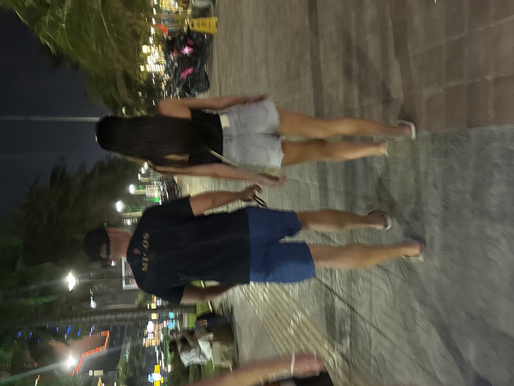A nighttime scene in Đà Nẵng, Vietnam, shows a couple walking along a paved promenade. The woman has long, dark hair and wears a black top with light-colored denim shorts. The man wears a black t-shirt, blue shorts, and a baseball cap. They are walking away from the camera, along the edge of a pathway lined with palm trees and illuminated streetlights. Distant city lights are visible in the background.