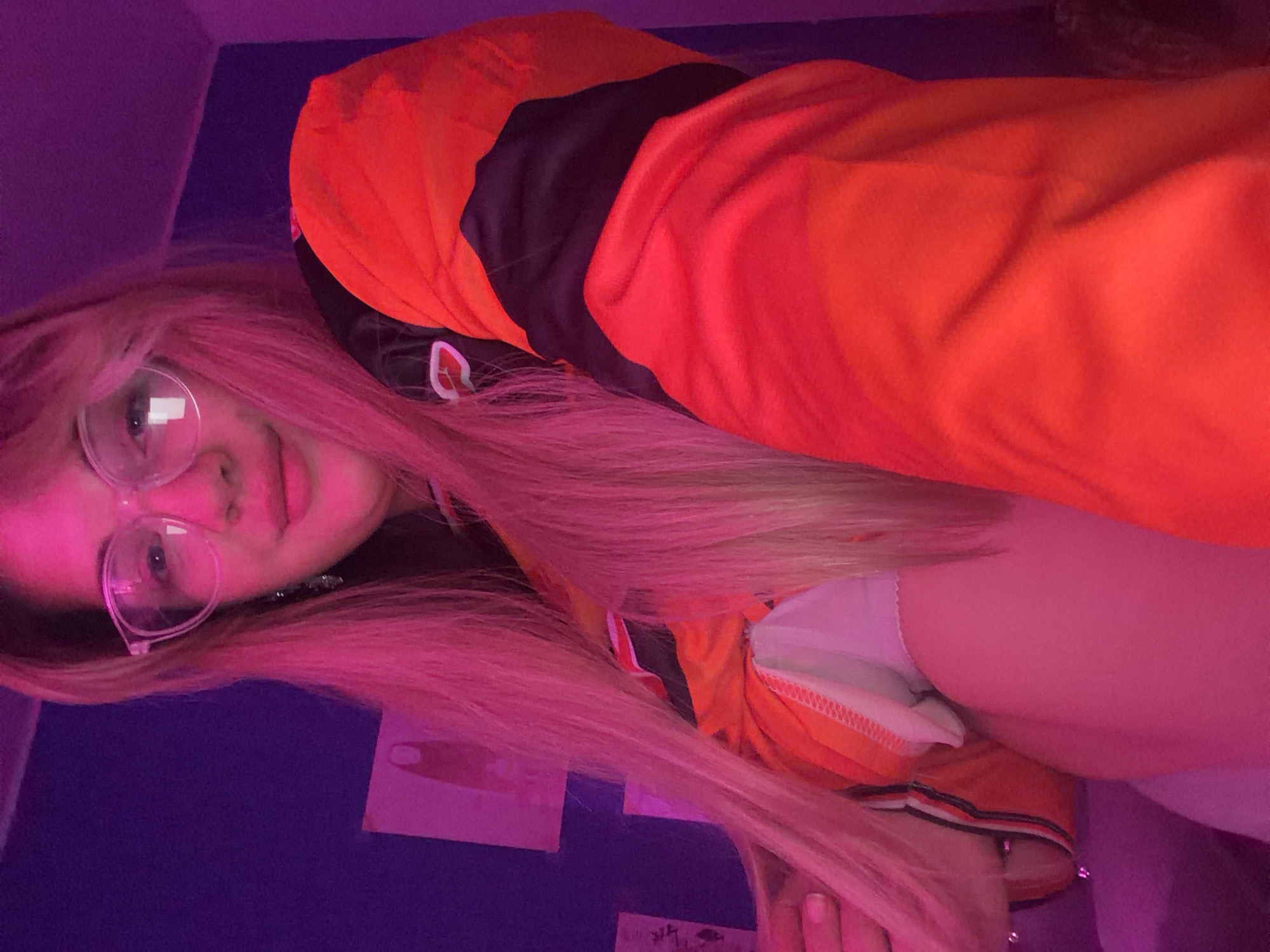 The image shows a young woman with pink hair, wearing glasses and an orange hoodie, lying on a purple surface. The location is not evident from the image itself and cannot be confirmed as being in Большая Шильна, Russia. There are some papers with writing on them behind her head.