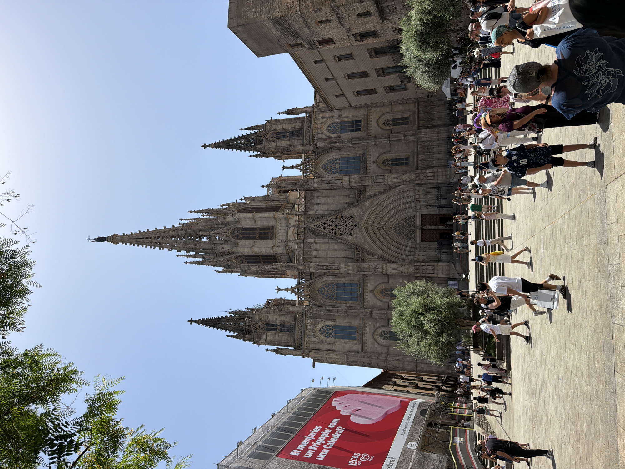 This is a low-angle, full-shot photograph of the Barcelona Cathedral in Barcelona, Spain, on a clear, sunny day. The cathedral's Gothic architecture, characterized by its spires and ornate facade, dominates the center of the frame. In the foreground, a crowd of tourists and locals are gathered on a paved plaza, some walking, some standing and talking, and some taking photos.
To the left of the cathedral, a large, red billboard with white text is attached to a building. The billboard features an image of a hand and text that appears to be an advertisement. The plaza is a light-colored stone, and in the middle ground, there are some trees. The sky is a clear blue. The scene is a typical daytime depiction of a popular tourist attraction in a European city.