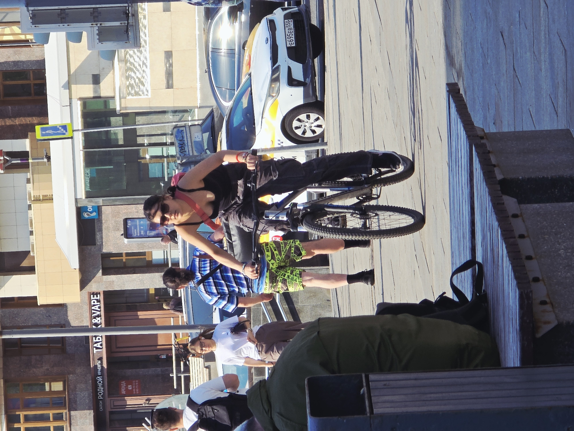 This outdoor shot in what appears to be Moscow, Russia, captures several pedestrians and a cyclist. A woman rides a bike along a sidewalk, adjacent to a row of people sitting on a bench. The scene shows urban life with buildings, shops, and vehicles forming the backdrop. One shop is labeled "ТAБ & VAPE" in Cyrillic script. A car with the license plate "E695EB 550" is parked nearby. The environment includes elements such as street signs, a crosswalk, and the sidewalk furniture. The subjects are engaged in various activities such as cycling, resting, and walking.