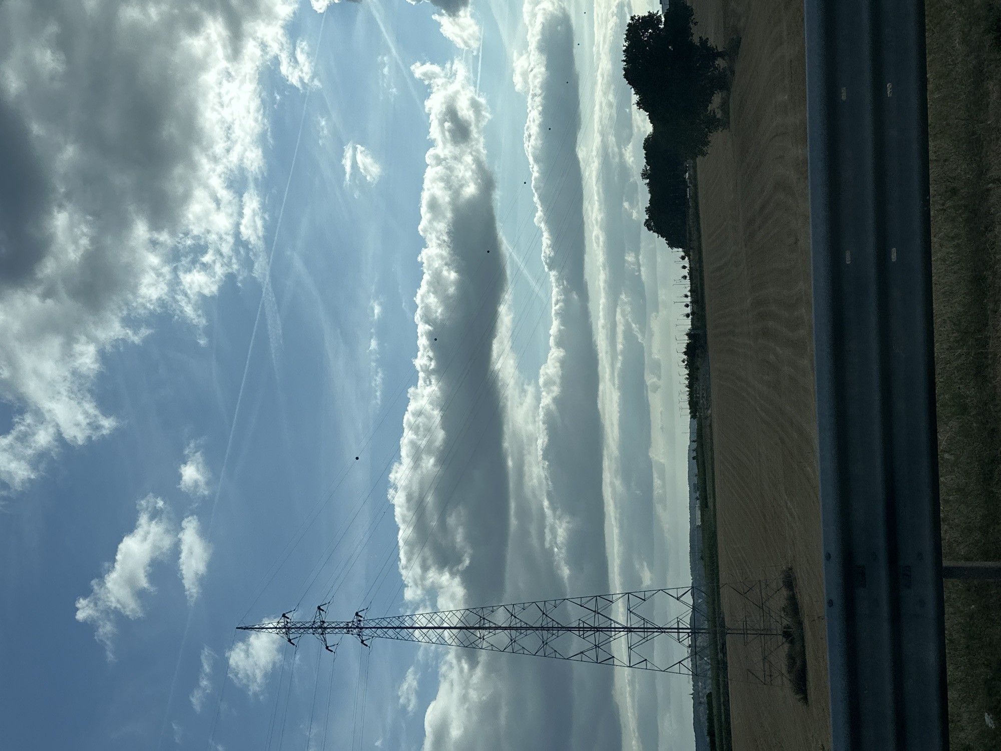 The image depicts a landscape under a cloudy sky, viewed from a roadside. In the foreground, a dark grey metal guardrail runs horizontally across the bottom of the frame. Beyond the guardrail, a field of dry, light brown grass or harvested crops stretches towards the horizon. A tall, lattice-like metal pylon, part of a power line, dominates the left side of the image. The pylon is set against a backdrop of layered clouds in a blue sky, with some contrails visible. A dark, rounded object, possibly a bird or a drone, is visible in the sky to the right of the pylon. In the distance, a line of trees and some distant structures, possibly buildings or wind turbines, are visible along the horizon. The overall impression is of a rural or semi-rural environment, likely viewed during daylight. The location is specified as the city of Namur, Belgium.