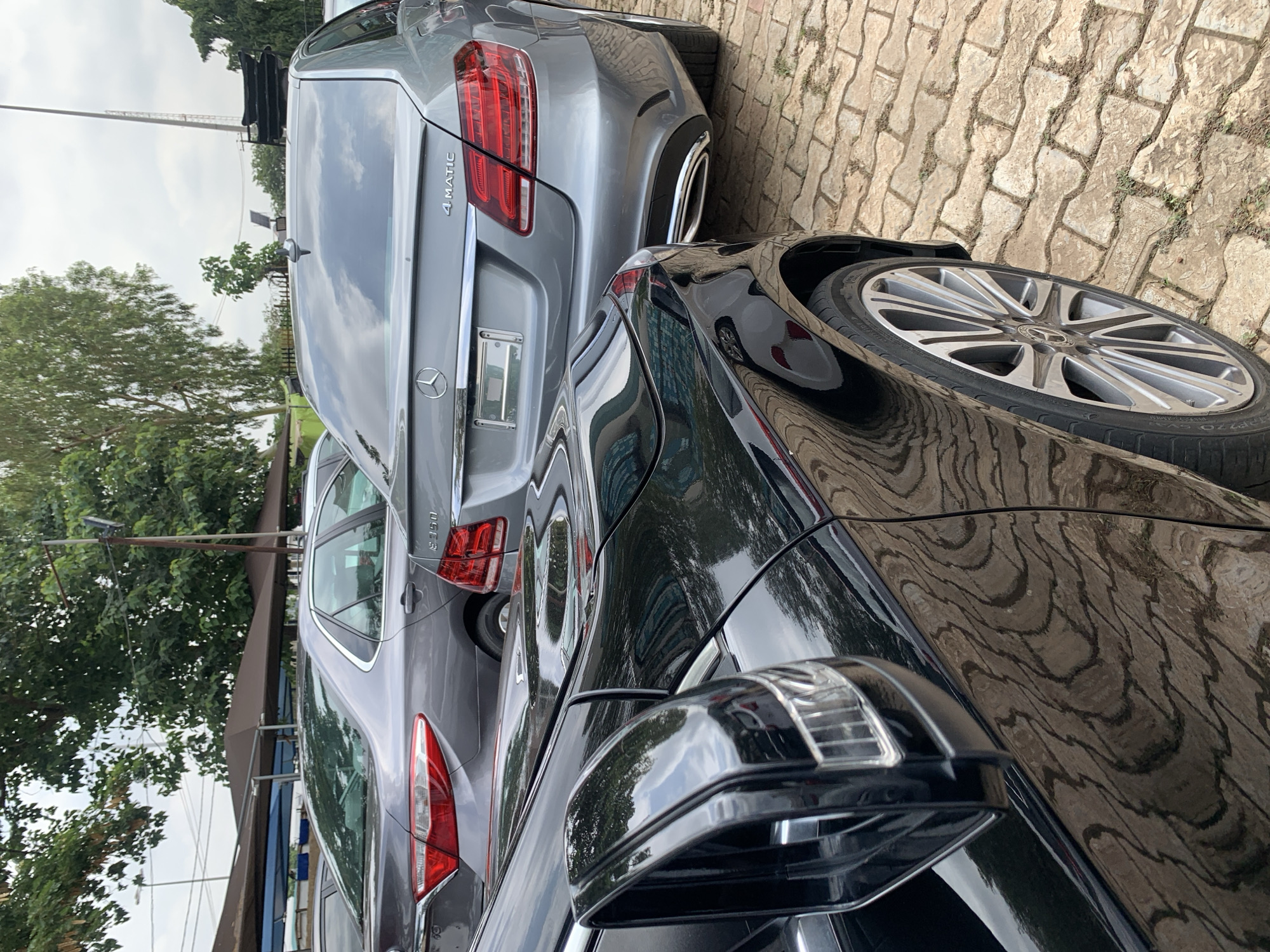 The image presents an outdoor scene, likely a car park or dealership in Abuja, Nigeria, showcasing two luxury sedans. The perspective is a low-angle shot, focusing on the vehicles parked on a paved surface.
In the foreground, a glossy black Mercedes-Benz sedan is positioned to the right. Its passenger side is visible, featuring a stylish multi-spoke alloy wheel with a prominent Mercedes-Benz logo at its hub. The tire bears the partially visible inscription "PIRELLI P ZERO." The car's highly reflective surface mirrors the intricate patterns of the light-colored interlocking pavers on the ground. A black side mirror with an integrated turn signal is visible in the bottom right corner.
Behind the black car, a grey Mercedes-Benz E-Class sedan is parked. Its rear section is clearly visible, displaying sleek red taillights, chrome trim, and a prominent exhaust outlet integrated into the bumper. The trunk lid features the "4 MATIC" badge, indicating its all-wheel-drive system, and the iconic Mercedes-Benz star emblem. On the side, the model designation "E350" is partially visible.
The ground beneath the cars is paved with light-brown, textured interlocking stones, forming a distinct, undulating pattern. The background consists of green trees and an overcast, cloudy sky, indicating daytime, likely late morning or afternoon. A tall, slender utility pole or light pole is visible in the distance. The scene is devoid of people, portraying a static arrangement of parked vehicles.