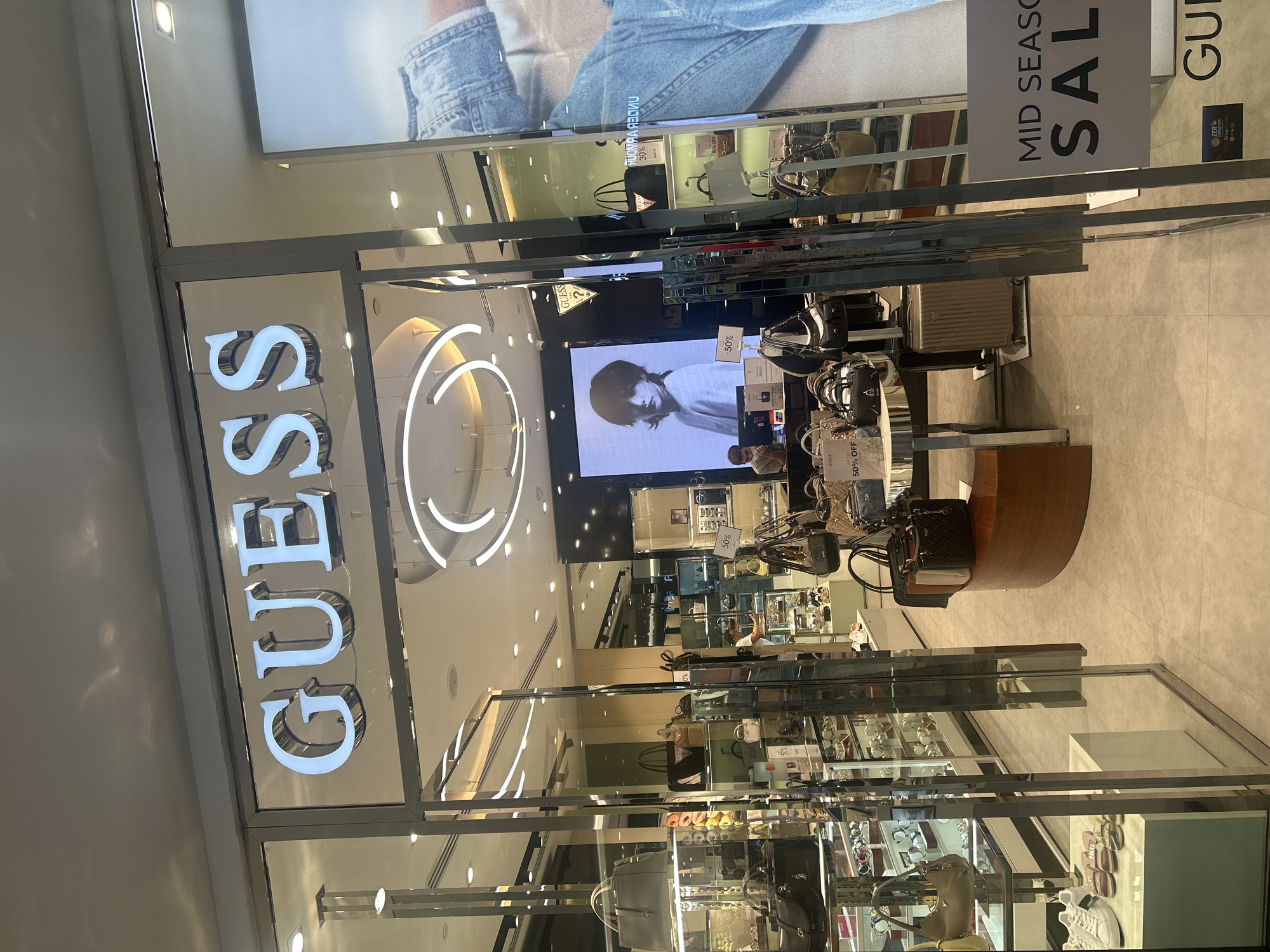 The image shows the front of a Guess store, likely inside a mall or shopping center in Patong, Thailand. The "GUESS" sign is vertically oriented on the left, with illuminated, metallic letters. A large, circular neon light fixture is visible beside the sign. A display screen shows a black and white image, possibly a model. Merchandise, including bags and accessories, is visible inside the store. Sales signs, including "MID SEASON SALE" and percentage discounts (50% off), are displayed. The interior of the store appears well-lit. The environment is clean and modern, with reflective glass surfaces.