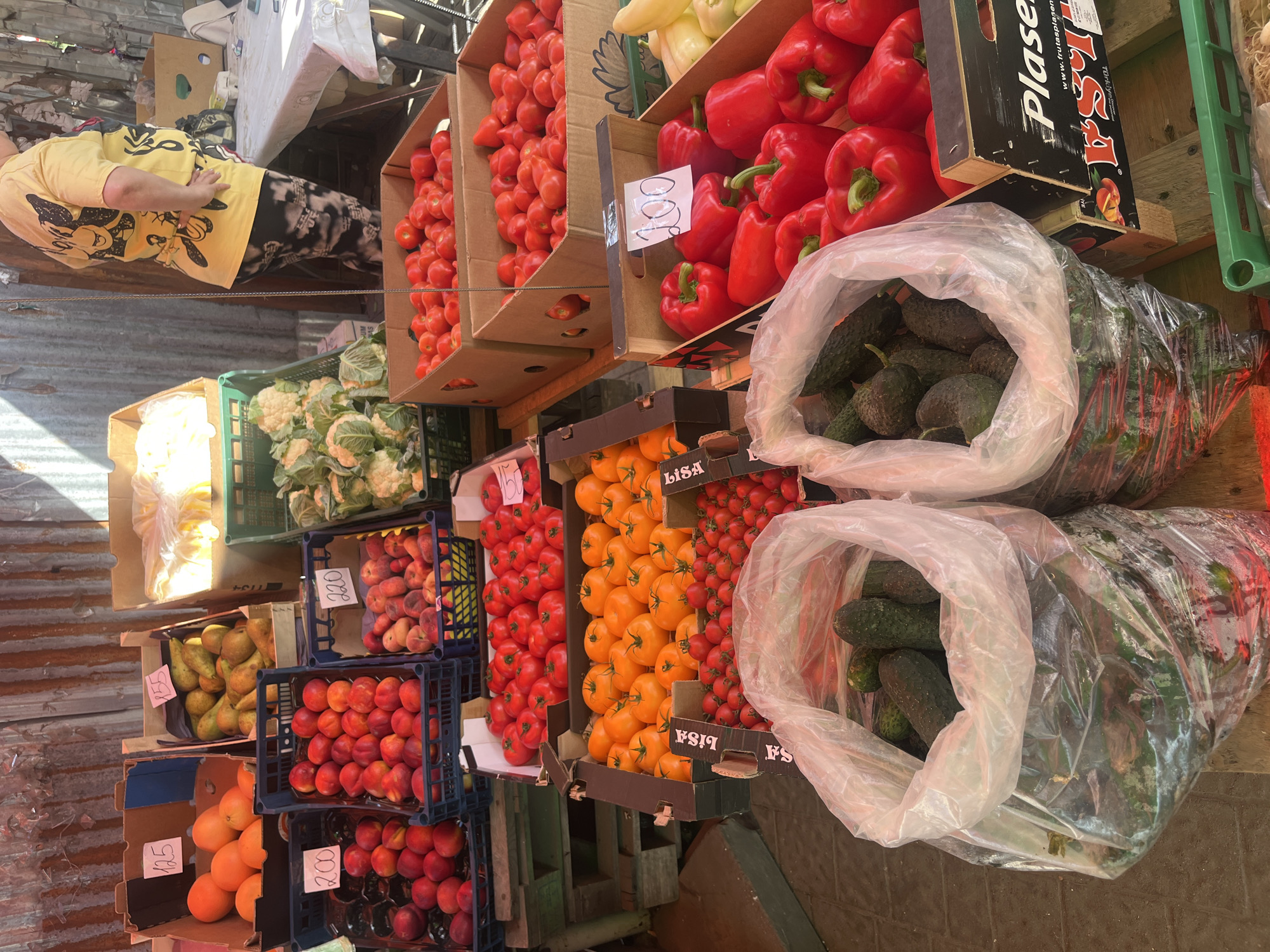 This is a medium shot of a fruit and vegetable market stall in Uzhhorod, Ukraine. The stall is outdoors, with a corrugated metal wall in the background. A woman in a yellow t-shirt and patterned leggings stands behind the stall. Various fruits and vegetables are displayed in boxes and crates, including oranges, pears, peaches, tomatoes, cauliflower, cucumbers, and red bell peppers. Some of the produce has price tags. The ground is paved with cobblestones. The scene appears to be illuminated by natural sunlight.