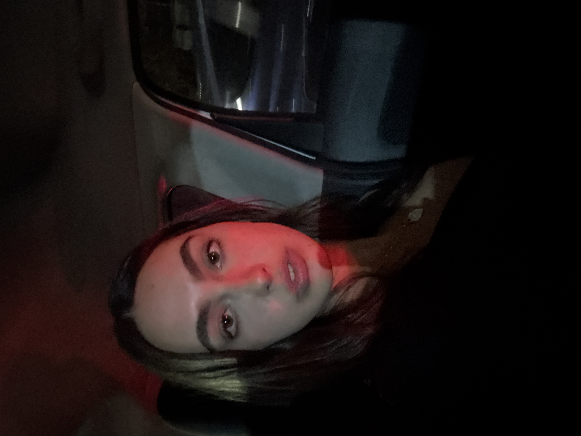 A medium shot features a young woman with light skin, dark brown hair, and brown eyes. She is positioned near a vehicle, seemingly sitting in a car, as indicated by the window in the upper portion of the frame. The car's interior is predominantly dark, and the woman is lit by a red light source, possibly from a traffic signal or a nearby light. The context implies the location might be Mexico City, Mexico, given the prompt's instructions. The focus is on the woman's face, which is turned slightly to the left. She appears to be looking directly at the camera. The environment is dark, with the red light casting a dramatic hue on her face.