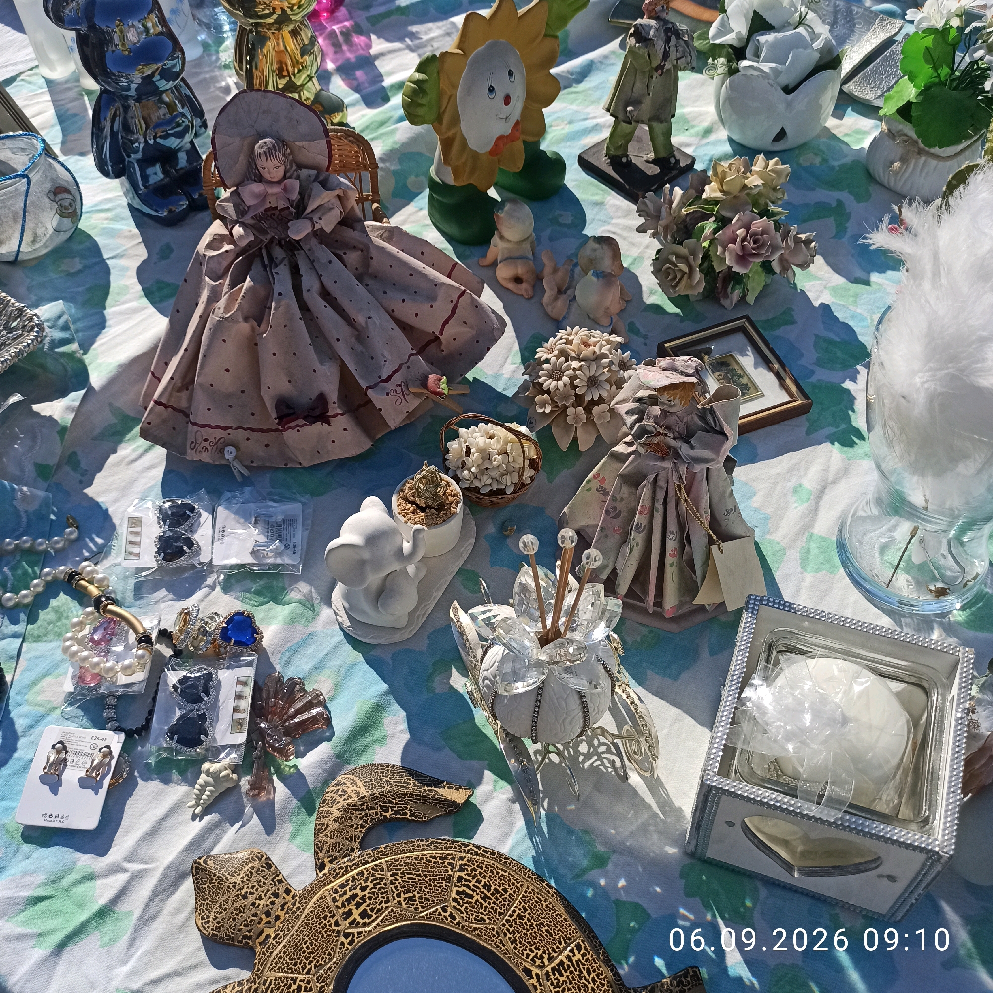 This is a close-up, overhead shot of a flea market stall in Sannicandro di Bari, Italy, on September 6, 2026, at 9:10 AM. The scene is laid out on a patterned tablecloth, showcasing a variety of trinkets and decorative items. Prominently featured are two porcelain dolls, one larger and seated in a wicker chair with a voluminous dress, and another smaller doll with a pale face and a floral dress. Various decorative floral arrangements, both real and artificial, are scattered throughout. Among the items are a ceramic sunflower figure with a painted face, a small white elephant figurine, a glass jar filled with white feathers, and a decorative golden turtle shell with a mirror. Several small boxes of jewelry, including earrings and rings with colorful stones, are also visible. In the foreground, a silver-colored box with a heart-shaped cut-out and a white object inside is present. The lighting suggests it is daytime, with distinct shadows cast by the objects.