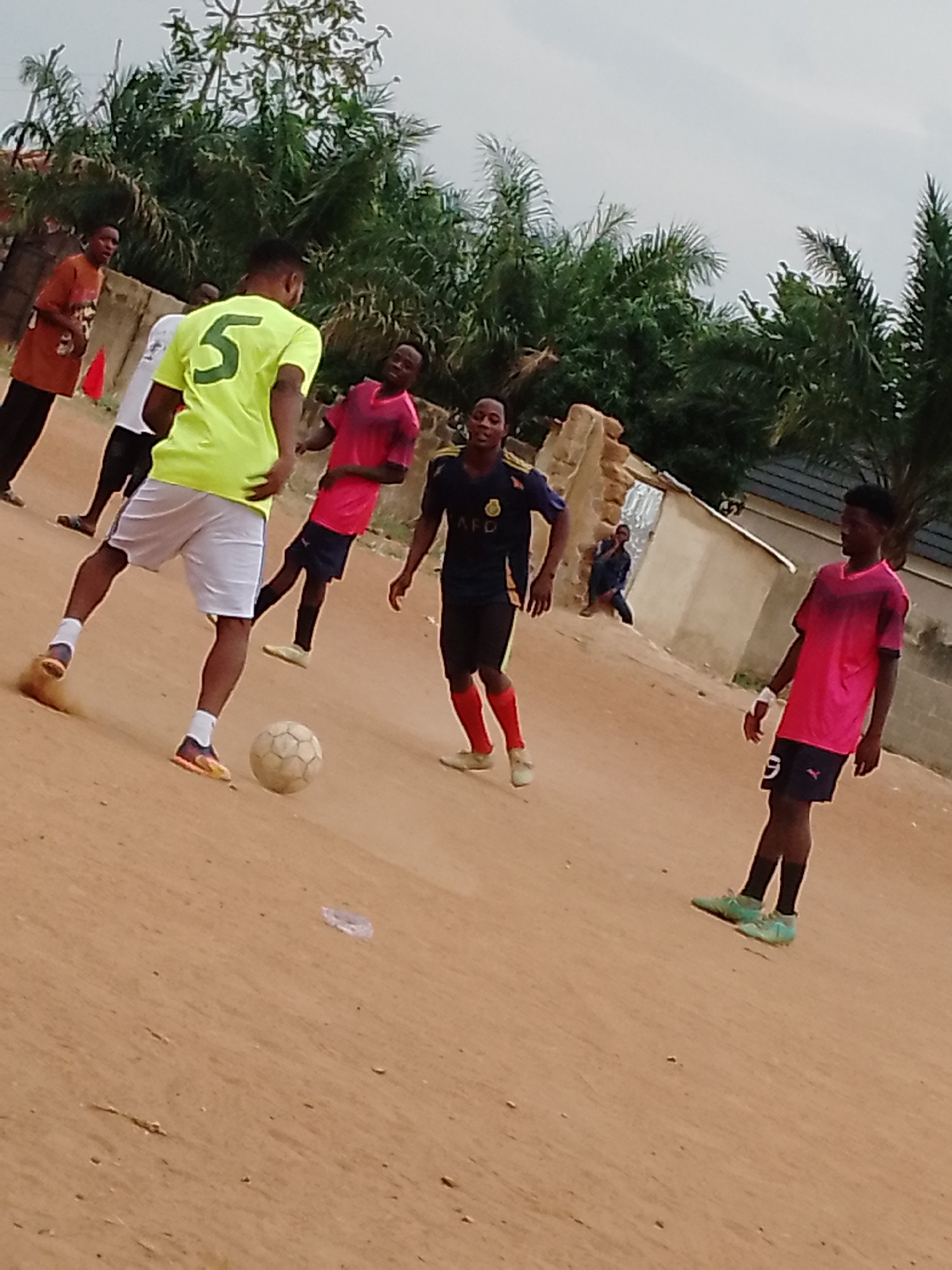 A street soccer match is in progress on a dirt field in an unidentified urban location. A player in a bright yellow jersey with the number "5" on the back is kicking a soccer ball. Several other individuals are nearby, including a player in a pink shirt with the number "9", a person in a dark blue jersey, and others dressed in casual attire. The background includes trees, a building, and a person sitting near the building. The sky is overcast.