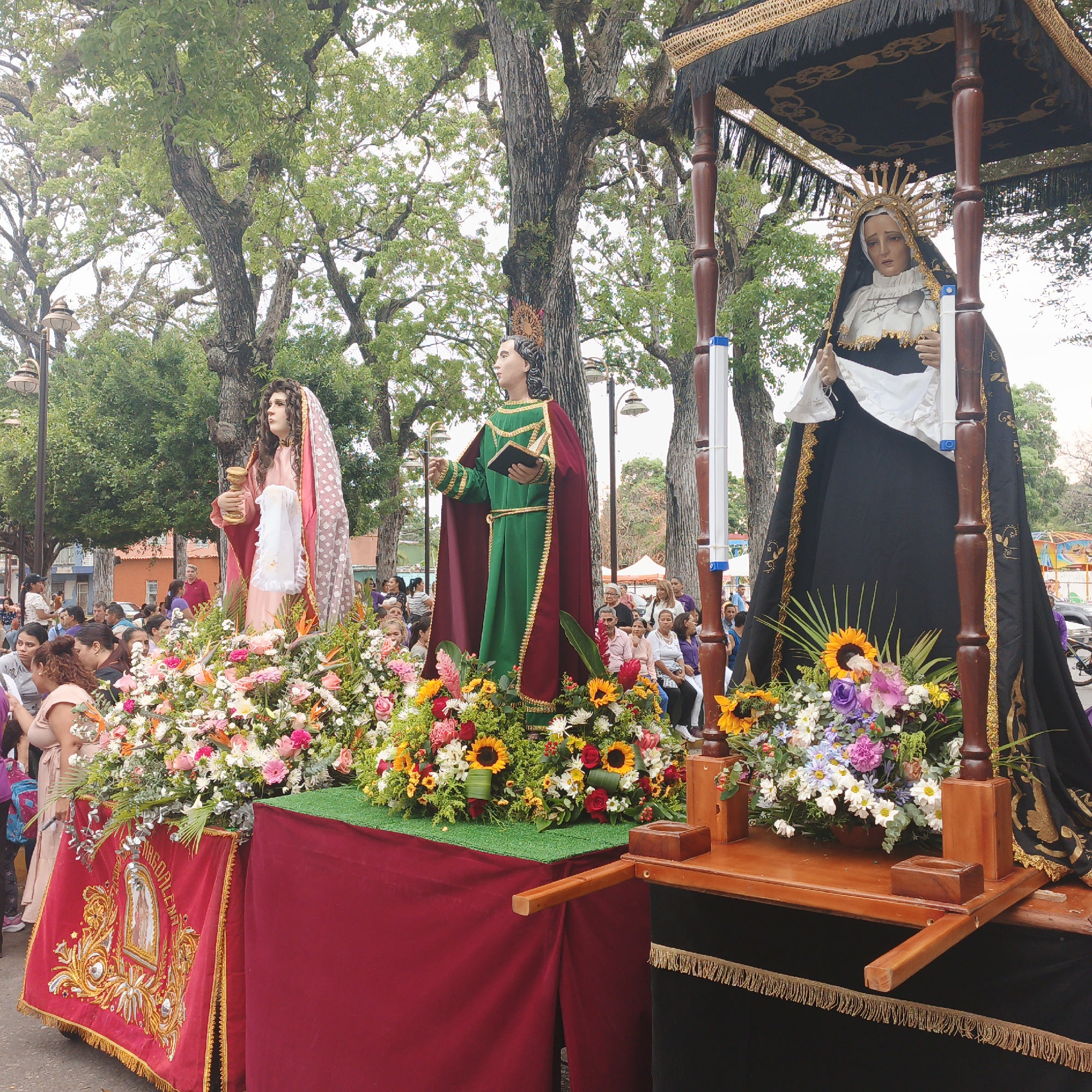 The image shows a religious procession in Tinaquillo, Venezuela. Three floats are visible, each carrying a statue. The first statue depicts a woman in a pink dress and veil, holding a golden chalice. The second statue is of a male figure in green and red robes, holding a book. The third depicts a woman in a black and white robe and a golden crown, standing under a black canopy. Each float is adorned with a variety of flowers. A crowd of people lines the street, observing the procession. Trees are visible in the background, suggesting the procession is taking place in a park or public square.