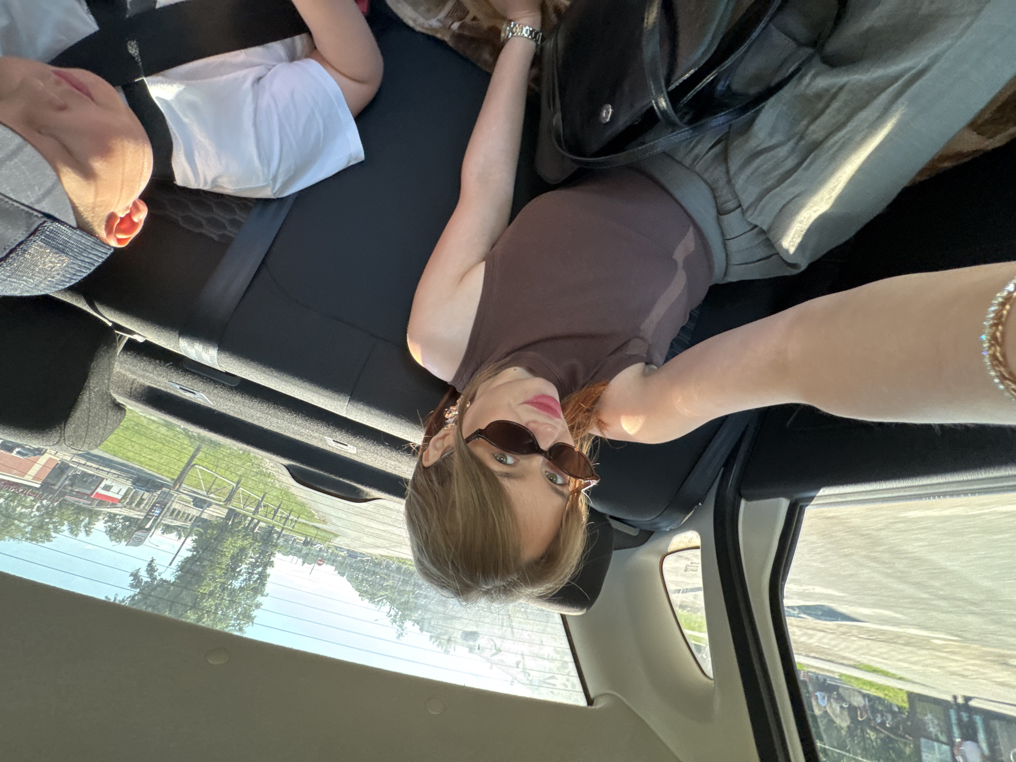 A woman and a young child are in the back seat of a car, which is driving on a street in Chaikovsky, Russia. The woman is taking a selfie. She is wearing sunglasses and a brown tank top, and a green skirt. The child is wearing a white t-shirt and a baseball cap. In the background, there are trees, buildings, and a fence. The sun is shining and it appears to be daytime.