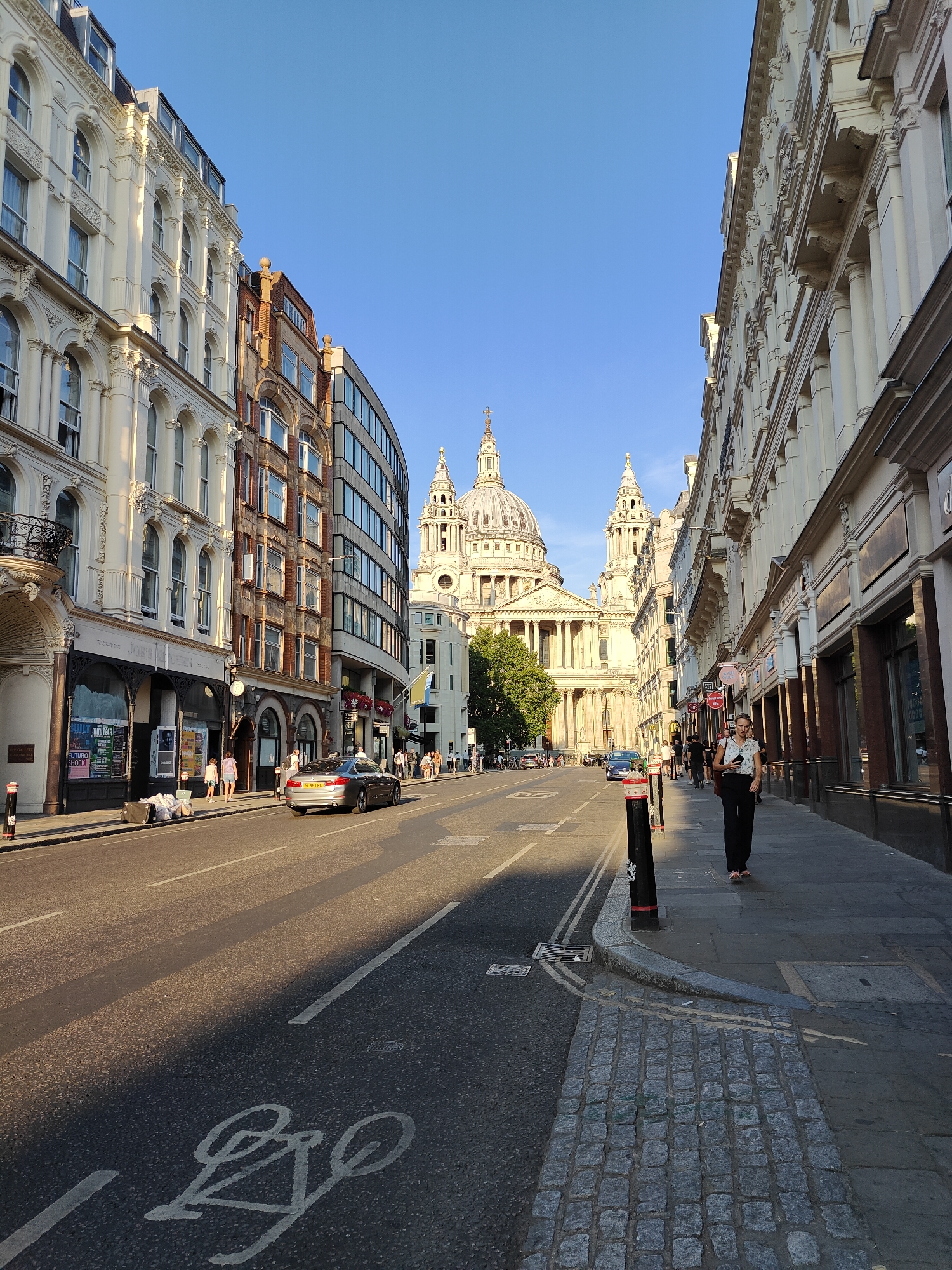 The image depicts a street scene in the city of London, United Kingdom, with St. Paul's Cathedral prominently featured in the background. The perspective is from street level, looking down a road lined with buildings on both sides.
On the left side of the street, there are several buildings. The closest building on the left has a white facade with ornate architectural details, arched windows, and a decorative balcony. Further down, there is a building with a red brick facade, also with arched windows, and next to it, a more modern building with a curved glass facade.
On the right side of the street, the buildings are also multi-story, with a more uniform, classical architectural style. These buildings have shopfronts at street level, many with large display windows.
The street itself is paved with asphalt and has visible lane markings, including a bicycle lane marking in the foreground. There is a silver car driving away from the camera in the middle lane.
Several people are visible on the sidewalks. On the left sidewalk, near the shopfronts, a few individuals are walking and standing. On the right sidewalk, a woman in dark trousers and a patterned top is walking towards the camera, carrying a bag. Further down the right sidewalk, more pedestrians are seen.
The environment is characterized by clear blue skies and the warm lighting of what appears to be late afternoon sun, casting long shadows across the street. The overall impression is of a busy urban environment in London.