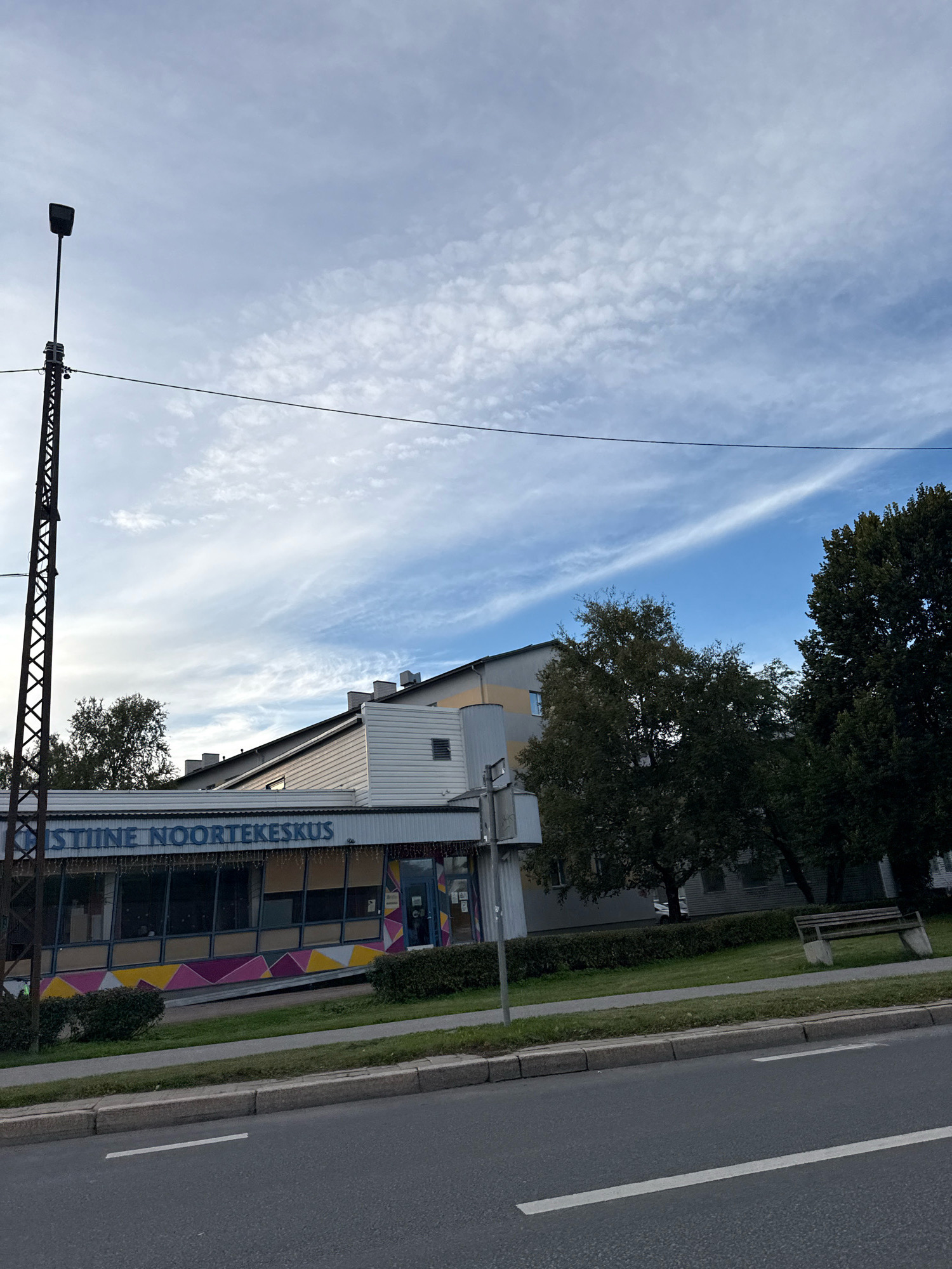 This is a photograph of a building with the text "KISTIINE NOORTEKESKUS" visible on its facade. The building appears to be a youth center, possibly located in Tallinn, Estonia. The facade features large windows with colorful geometric patterns below them. A street lamp stands to the left of the building, and trees and a sidewalk are also visible. The sky is cloudy with streaks of blue visible. The photograph is taken from across a street, with the road and a curb in the foreground. There is a bench visible on the grass area in front of the building.