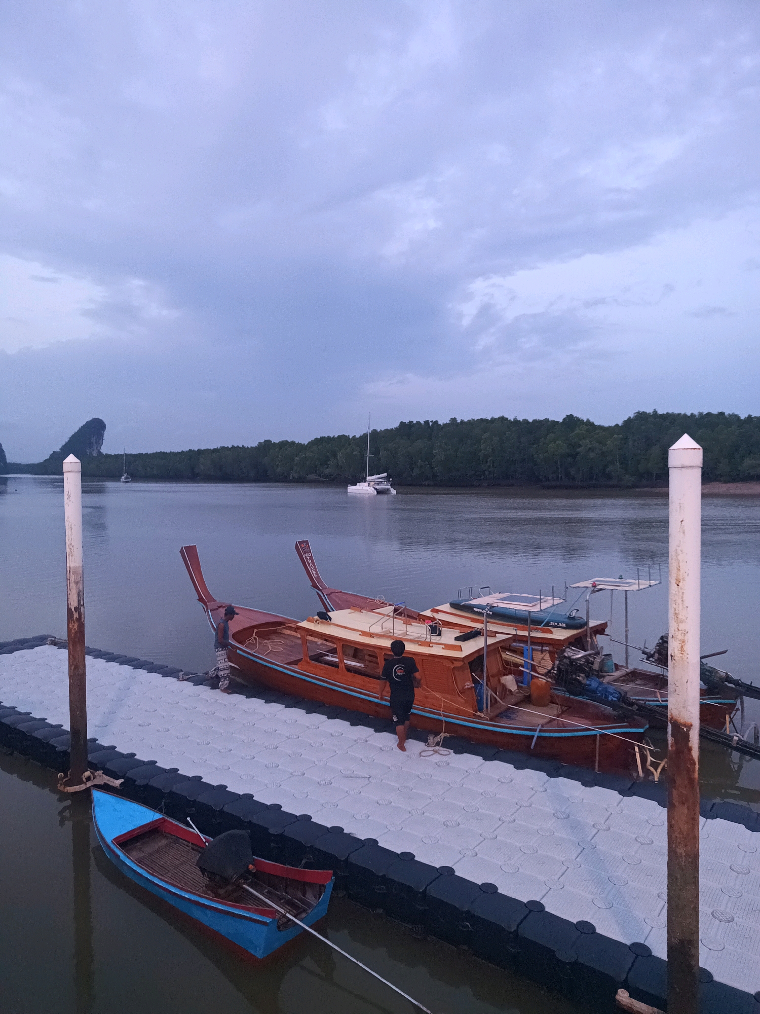 A vertical, eye-level, wide shot captures a serene dusk scene at a floating dock in Tambon Pak Nam, Thailand. The sky is overcast with soft grey and white clouds. A calm body of water, likely a river or bay, occupies the majority of the frame, reflecting the muted light.
In the foreground, a modular floating dock, constructed from interlocking grey plastic sections with a dark border, extends into the water. A single, weathered wooden post with a white cylindrical cap stands at the left edge of the dock. Attached to this post is a dark, weathered metal bracket.
Moored to the dock are several traditional Thai longtail boats, distinguished by their slender wooden hulls and distinctive front and rear extensions. The boats are predominantly wooden with a reddish-brown exterior, accented with light blue trim. One of these boats has a person, appearing to be a man wearing a dark t-shirt and patterned shorts, standing on it. He is looking downwards towards the boat. Another longtail boat is moored further back, its wooden structure visible.
To the left of the dock, a small, vibrant blue and red dinghy is tied to a mooring rope. A dark engine is mounted on the stern of this small boat.
In the middle ground, the tranquil water leads to a dense, dark green tree line along the shore, suggesting a mangrove forest or jungle. In the distance, a distinctive, jagged limestone karst formation rises from the water on the left. Several sailboats, one a catamaran and another a monohull, are visible on the water in the middle to background, their white sails contrasting with the muted environment.
The overall impression is one of peaceful twilight in a coastal or riverine setting.