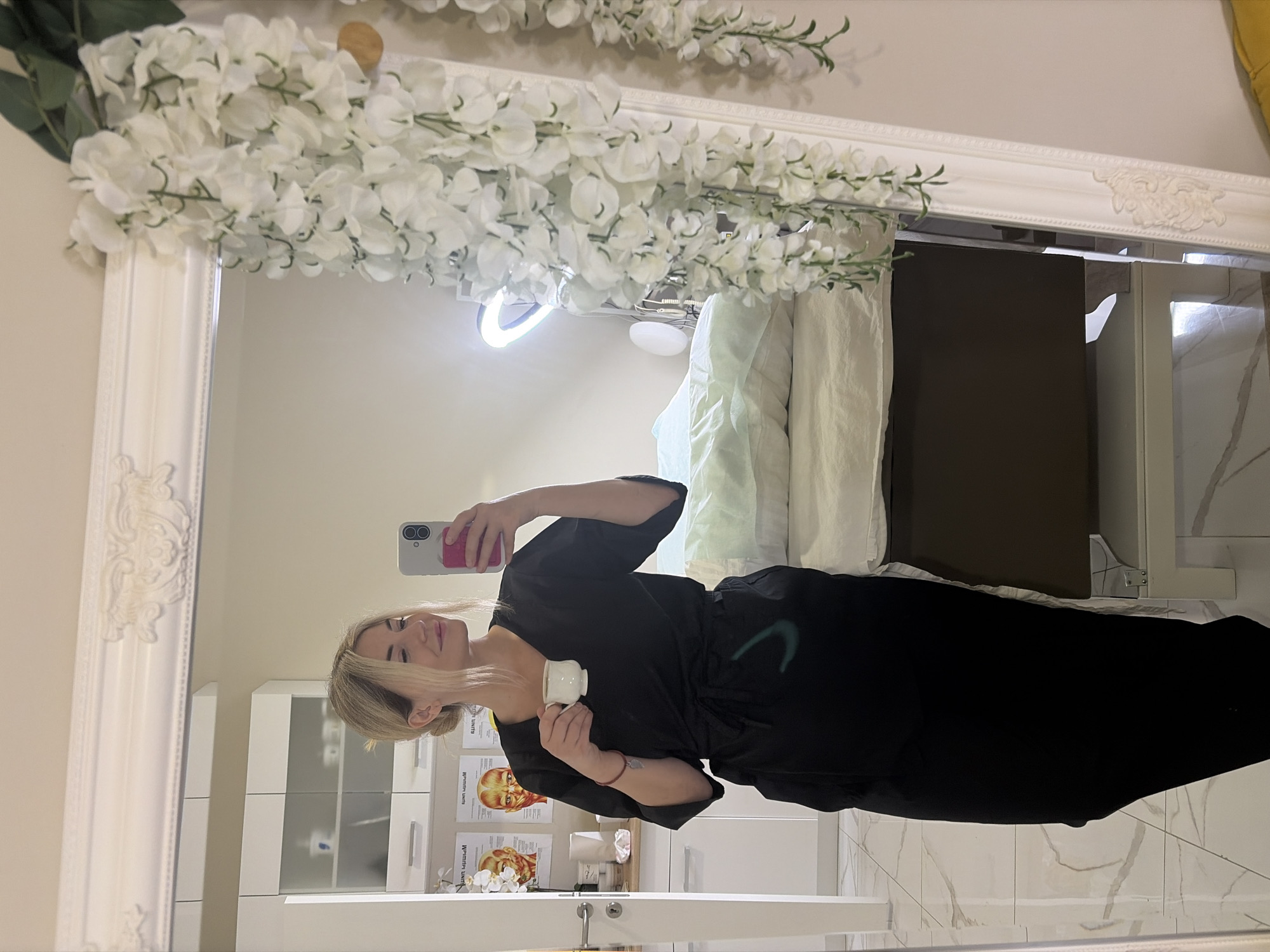 A woman is taking a mirror selfie in a brightly lit room, likely a spa or medical office. She is wearing a black scrub suit and holding a white cup with a beverage. Her blonde hair is pulled back in a bun. Behind her, a massage table is covered with white and light green linens. The room features white cabinetry, anatomical charts on the wall, and a ring light. The mirror is framed with ornate white molding and decorated with hanging white flowers. The floor is tiled with a marble pattern. The location is identified as Budva, Montenegro.