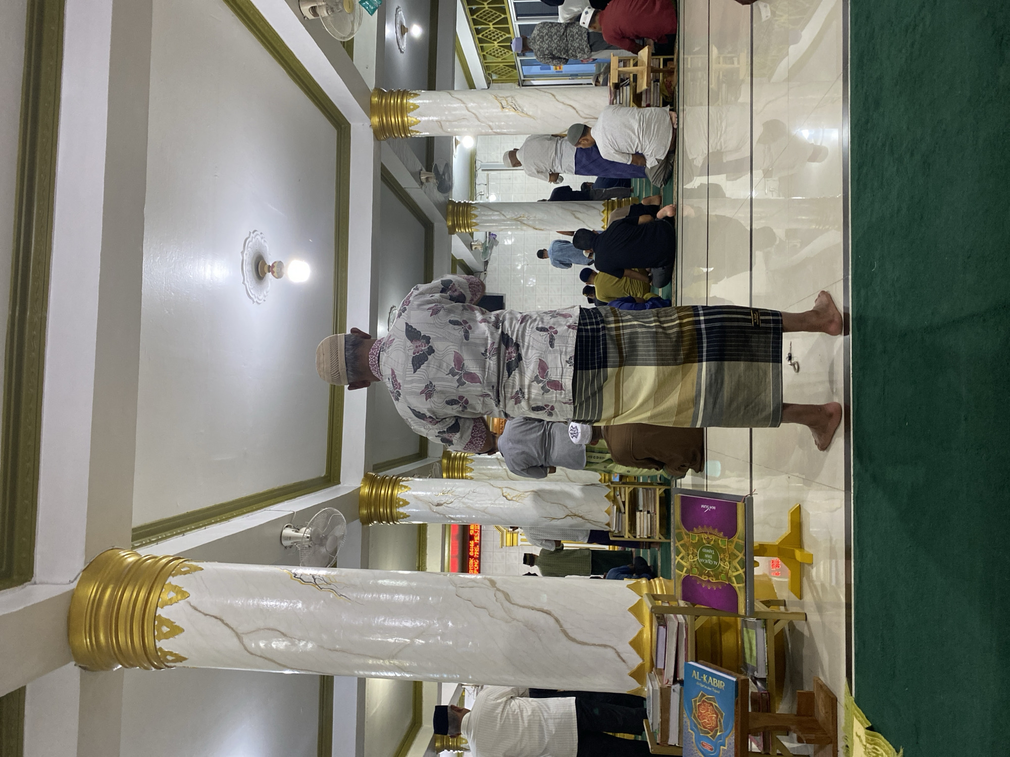 The image depicts the interior of a mosque in Lhokseumawe, Indonesia, during a prayer session. Several men are present, standing and kneeling on a green carpeted floor, facing the qibla. In the foreground, a man from behind is wearing a patterned shirt and a sarong. Large, white, marble-patterned pillars with gold accents line the space, supporting a white ceiling with ornate lighting fixtures and ceiling fans. Books are neatly stacked on shelves to the left of the foreground. The overall atmosphere is one of communal worship and reverence.