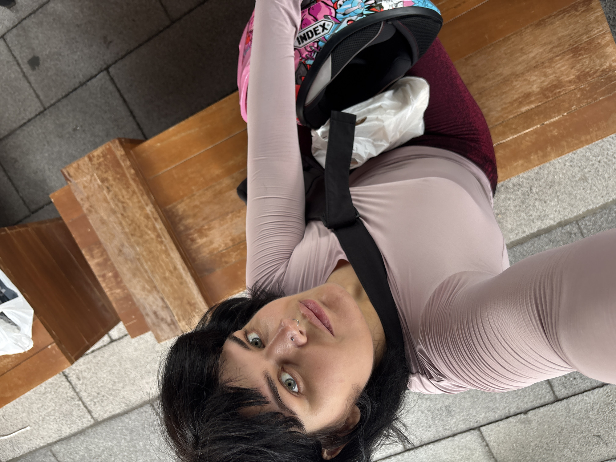 A person with black hair and light green eyes, wearing a pink long-sleeved shirt and maroon leggings, is sitting on a wooden bench. A black strap is visible across their chest, and a bag with colorful graphics is hanging from it. The person has a nose ring and a lip ring. The background shows a gray tiled walkway and a wooden bench with multiple levels. The scene is outdoors, and the lighting suggests it is daytime. The location is specified as Nong Pa Khrang, Thailand.