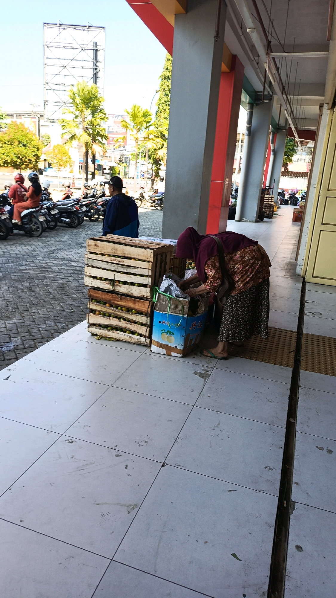 An outdoor market scene unfolds on a paved walkway adjacent to a building with red and gray pillars. In the foreground, two wooden crates filled with fruit, possibly oranges, are stacked. Beside the crates, a woman wearing a burgundy headscarf and a floral patterned top is bent over, interacting with a cardboard box. Her dark floral patterned skirt and green flip-flops are visible. To the left of the crates, a man wearing a dark blue and black hoodie stands facing away from the camera. In the background, several motorcycles are parked, and on one of them, two people are seated, both wearing helmets. The environment suggests a sunny day, with tall palm trees and a billboard visible in the distance. The overall scene depicts a busy marketplace in Nologaten, Indonesia.