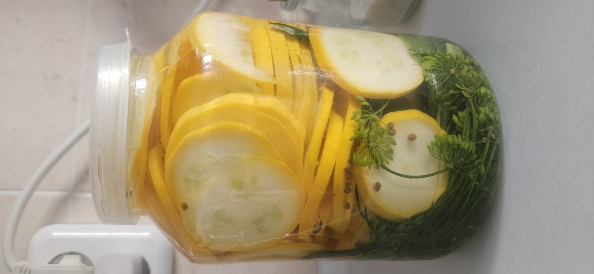 A clear glass jar filled with sliced yellow squash and dill pickles sits on a white countertop. The jar is sealed with a white plastic lid. In the background, a white electrical outlet and cord are visible on a beige tiled wall. The lighting is bright and evenly distributed. There are no people present in the image. The image appears to be taken in a kitchen or pantry. The location is a domestic setting, possibly a home kitchen in Vitsebsk, Belarus.
