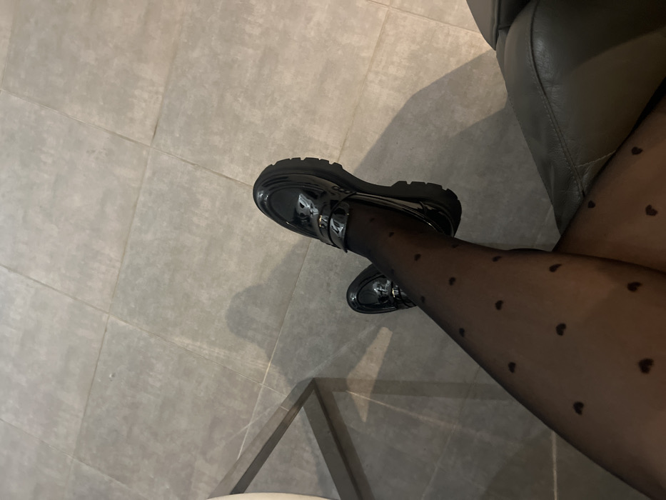 A person wearing black tights with small heart patterns is seated. The legs are crossed at the knees. The person is wearing black shiny penny loafers with thick soles. The background consists of gray floor tiles and a portion of a gray leather chair or couch to the right. The lighting casts shadows.