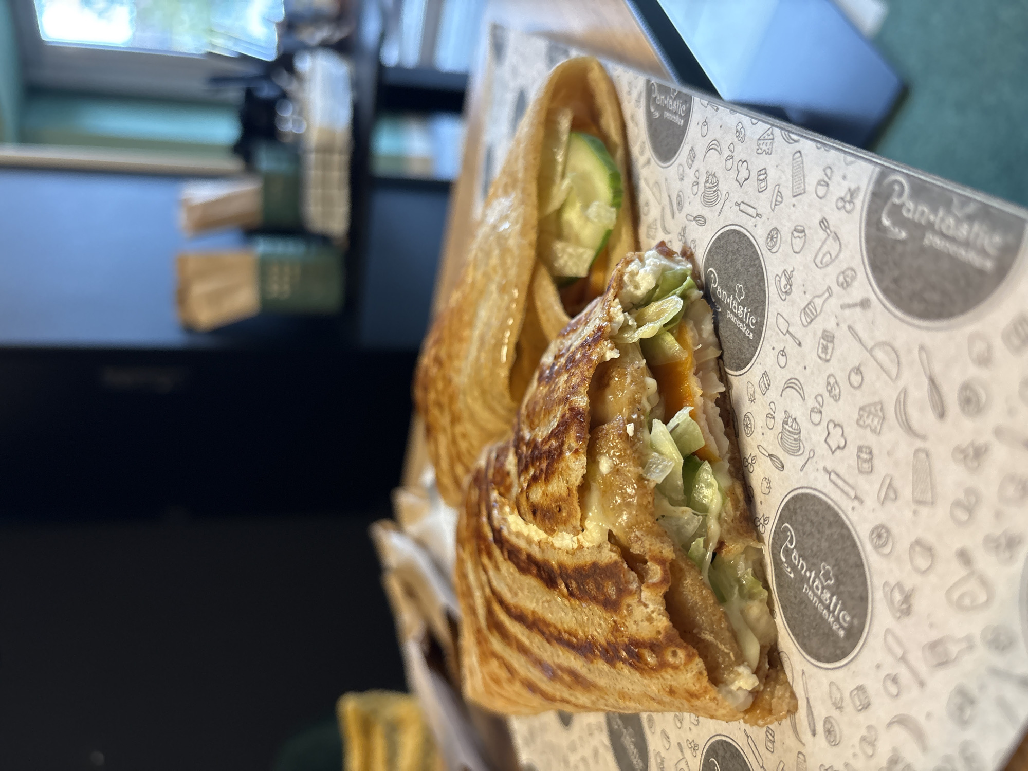 The image shows a partially eaten savory crepe or pancake in Veliko Tarnovo, Bulgaria. The pancake is filled with lettuce, cucumber, and other ingredients. It's served in a branded "Pan-tastic Pancakes" paper container. The background shows parts of a table and other blurred objects.