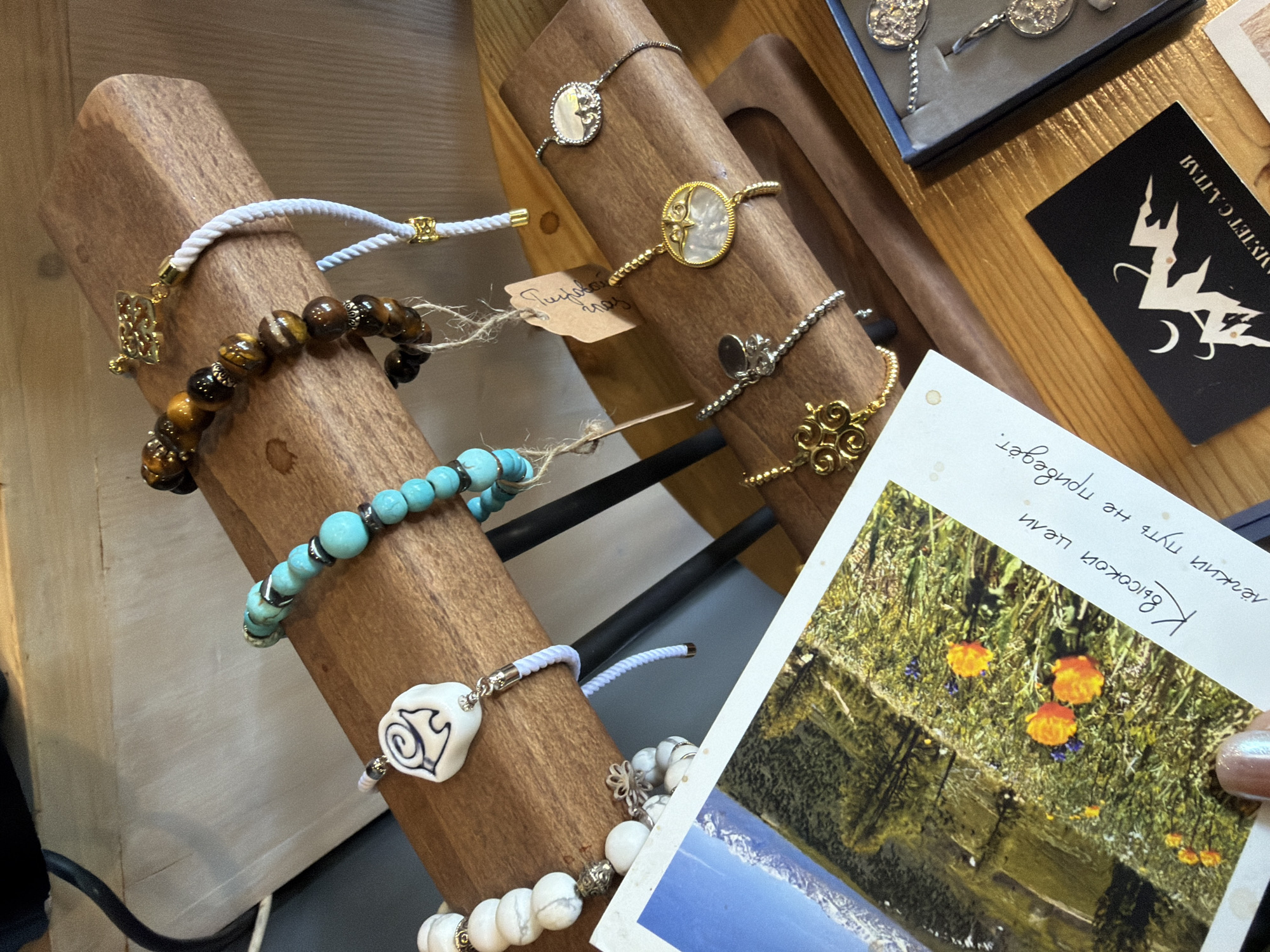 The image shows a display of bracelets at a shop in Mayma, Russia. The bracelets are arranged on wooden stands. There are bracelets with different types of beads including white, blue, brown, and tiger eye. Some bracelets have charms. A small box is visible to the right, containing more silver jewelry. A business card with a logo in the upper right corner states "Алтай. C Алтая". A postcard of a landscape with orange flowers and text is in the foreground to the right. A manicured hand is seen holding the postcard. The shop appears to be on a wooden table.