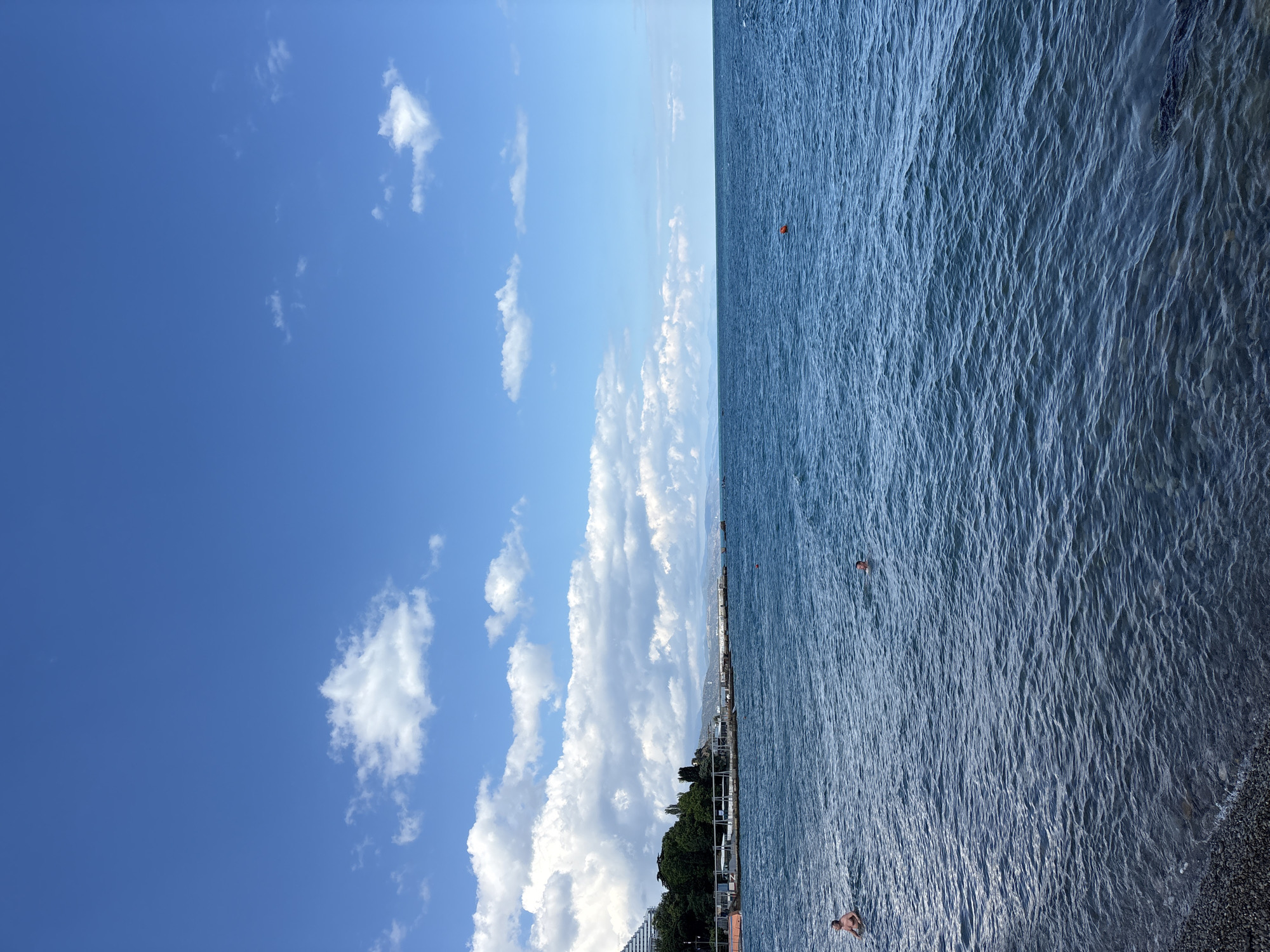 The image depicts a coastal scene in Alushta, Ukraine, on a clear day with a blue sky dotted with white clouds. The foreground features a pebbled beach transitioning into the rippling blue water of the sea. Two individuals are visible in the water, one closer to the shore and another further out, both appearing to be swimming or bathing. In the midground, a coastline is lined with lush green trees and buildings, including a large white structure that resembles a hotel or resort. A pier or promenade extends into the water, with several small boats or buoys visible. The horizon is defined by the distant outline of hills or mountains under the expansive sky. A single orange buoy is present in the water in the middle right portion of the frame.