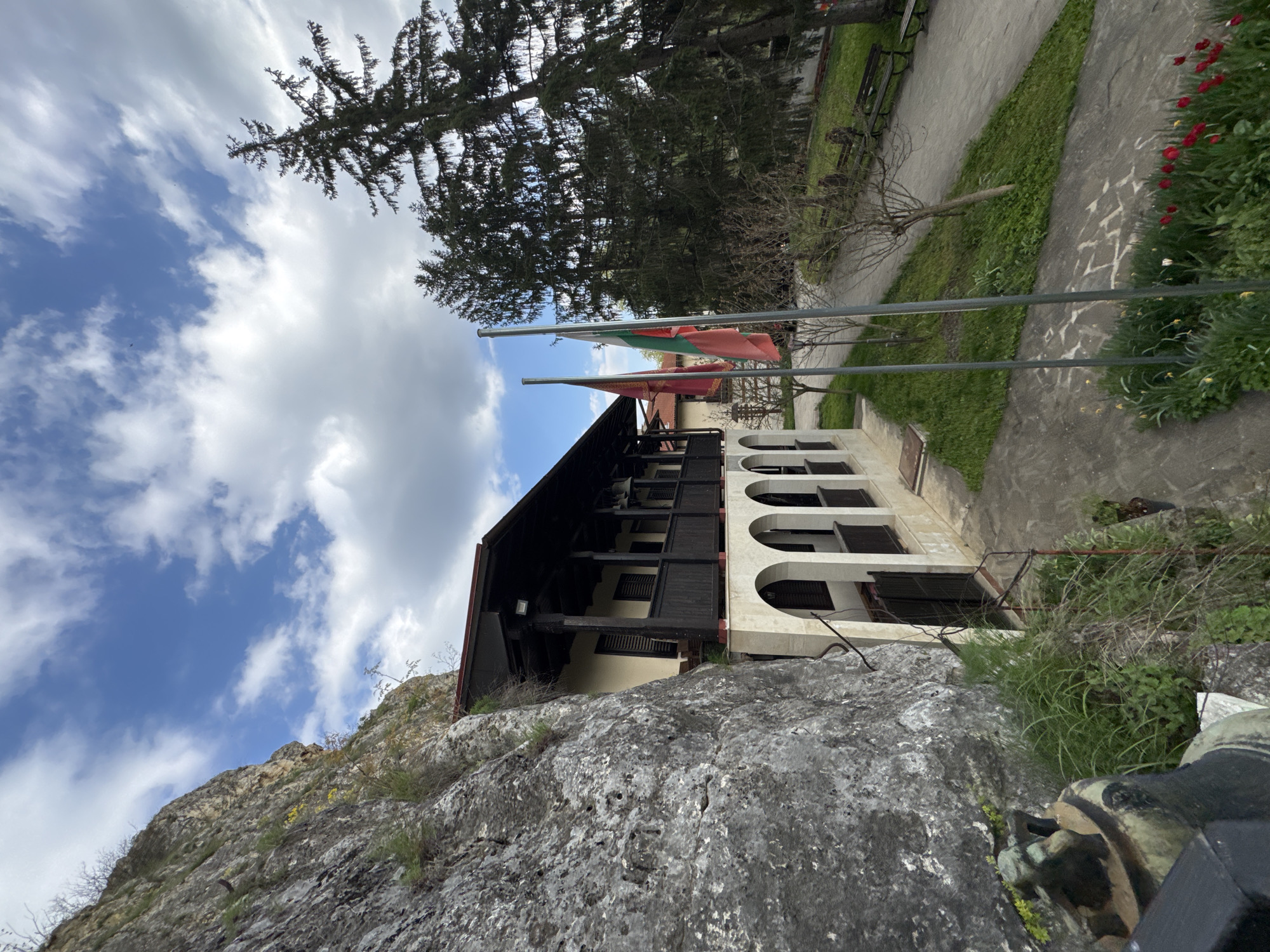 The image depicts a multi-story building with traditional architectural elements, possibly a monastery or historic house, situated adjacent to a rocky cliff face. The building features a dark, possibly wooden, upper section with enclosed balconies or verandas, supported by a lighter-colored lower section with arched openings. Two flags, presumably national or regional banners, are hoisted on poles near the building. A paved walkway or path runs alongside the building, bordered by greenery, including grass and flowering plants. In the distance, a bench and possibly other park-like features can be seen. The sky is partially covered with clouds. The composition suggests that the photograph was taken from a lower vantage point, perhaps near the base of the cliff, with the lens tilted upwards. The location is likely in or near Basarbovo, Bulgaria.