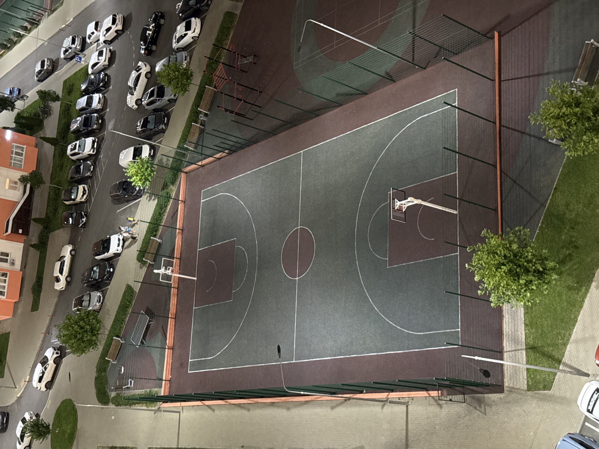 This is an aerial, daytime, medium-shot photograph of an outdoor basketball court and surrounding area in Anapa, Russia. The basketball court is the central focus, with a dark gray playing surface and maroon out-of-bounds areas. White lines mark the boundaries and key areas of the court, including a center circle, free-throw lines, and three-point arcs. A basketball hoop and backboard are visible on one end of the court.
To the left of the basketball court is a paved pathway or sidewalk. To the right and behind the court is a fenced-off area containing a small outdoor gym with red bars and a wooden bench, and more seating in the form of wooden benches. Beyond the gym area, there are more benches and a grassy area with trees.
A parking lot with numerous cars is visible in the upper portion of the image. The cars are of various colors, including white, black, and silver. Some of the cars are parked in designated spots. In the background, there are buildings, including one with an orange facade and windows. A street light is visible near the basketball court. There are a few people, appearing to be two children and an adult, visible near the edge of the parking lot, close to the basketball court.