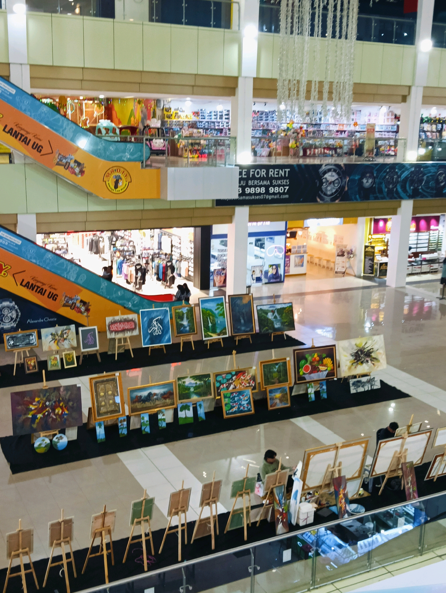This image depicts an indoor art exhibition or market located in Batam, Indonesia. The scene is set within a multi-level shopping mall, with an escalator visible in the upper left corner bearing the text "LANTAI UG" (which translates to "Ground Floor" in Indonesian). The exhibition features numerous paintings displayed on easels, arranged in rows on the mall's floor. The paintings vary in subject matter, including landscapes, abstract art, and still lifes, with some appearing to be for sale. In the lower part of the image, two individuals are visible actively painting at easels, suggesting a live art demonstration or artists at work. The environment is well-lit, characteristic of a shopping mall interior, with polished floors reflecting the ambient light. Stores with merchandise are visible on the upper levels of the mall, indicating a bustling commercial setting.