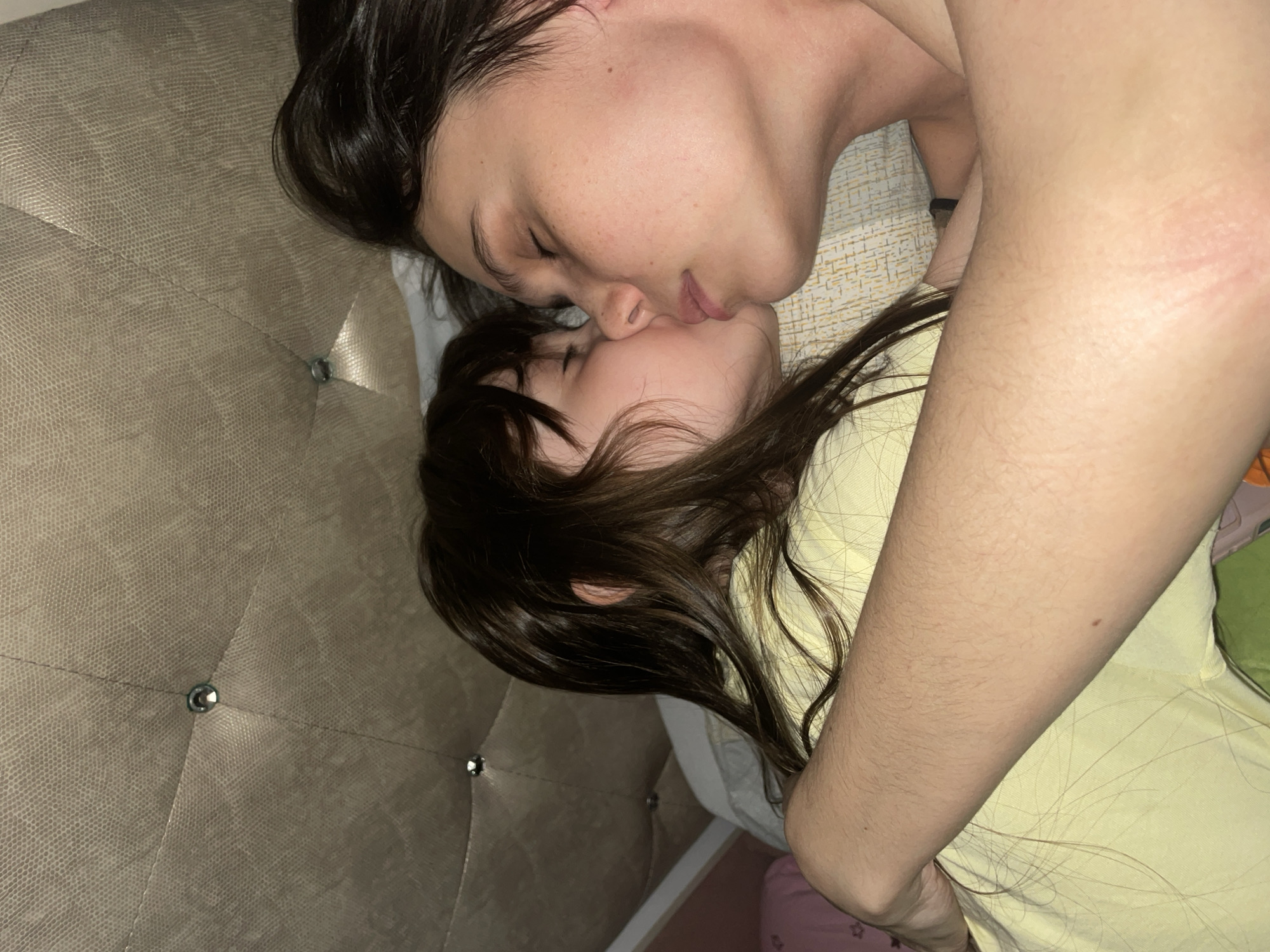 A top-down medium shot depicts an adult and a child lying on a bed with a light-colored, textured headboard. They are face-to-face and their lips are touching. Both have dark hair. The adult is closer to the camera and has fair skin. The child is partially covered by the adult's arm, which is also fair-skinned with noticeable hair. The child is wearing a yellow garment. A portion of bedding in various colors is visible. The location is unknown and is not discernible from the image.