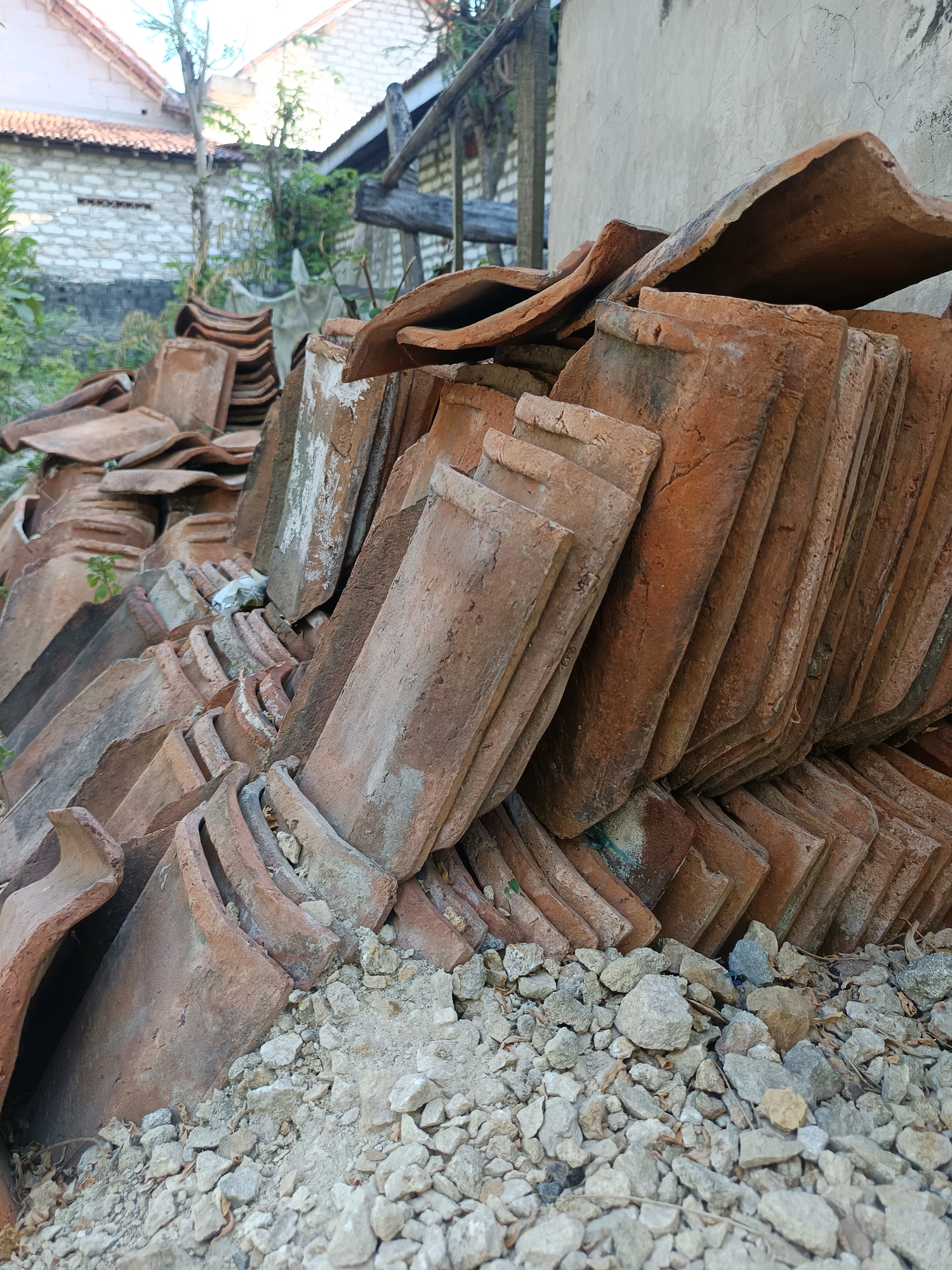 A pile of weathered, terracotta roof tiles is stacked haphazardly in an outdoor area. The tiles vary in condition, with some appearing cracked and chipped, while others retain their original curvature. They are predominantly reddish-brown in color, showing signs of age and exposure to the elements. The foreground consists of a layer of small, light-colored stones and some dried leaves. In the background, a portion of a building with a white facade is visible, along with some green foliage and a wooden structure. The scene suggests a construction or demolition site, possibly in a residential area of Gunung Sekar, Indonesia, given the architectural style hints. There are no people or animals present in the image.