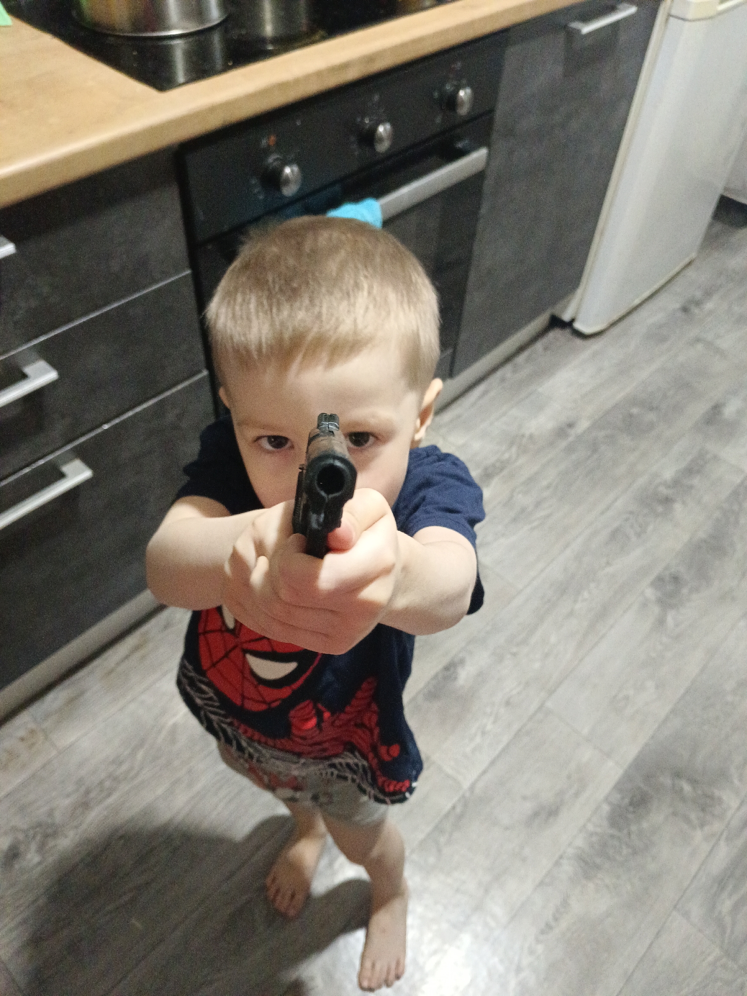 The image captures a young child, approximately 2-4 years old, standing barefoot on a light grey plank floor in a modern kitchen, located in the city of Chaikovsky, Russia. The child, with light blonde hair and wearing a dark blue t-shirt adorned with a red Spider-Man logo and light grey shorts, holds a black object resembling a handgun with both hands, aiming it directly forward towards the viewer. The child's gaze is fixed on the camera.
In the background, a dark grey kitchen cabinetry system with silver horizontal handles is visible. A light brown countertop extends above the cabinetry, supporting a black ceramic cooktop with a silver pot on its left burner. An integrated black oven with silver control knobs is situated within the cabinetry beneath the cooktop. To the far right, a partial view of a light-colored refrigerator is present. The environment appears to be an interior space, well-lit.