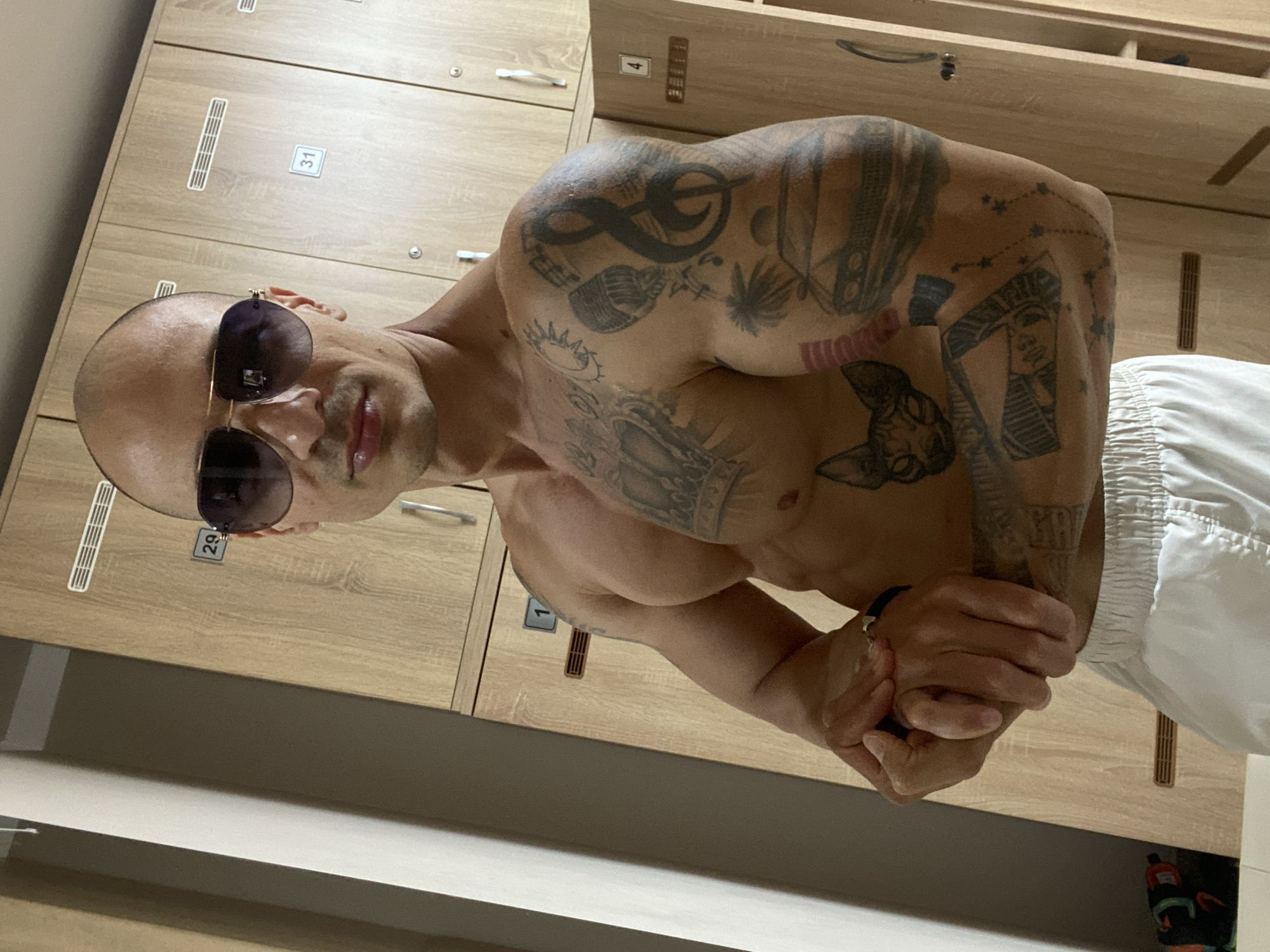 The image shows a muscular man with a shaved head and sunglasses posing in a locker room. He has several tattoos on his chest and arms, including a crown, a treble clef, a cat, and what appears to be a car. The locker room has wooden lockers with numbers on them, such as 29, 31, 1, and 4. The man is wearing white shorts. The lighting suggests an indoor setting. There is no information in the image to indicate the location in the city of Pleven, Bulgaria.