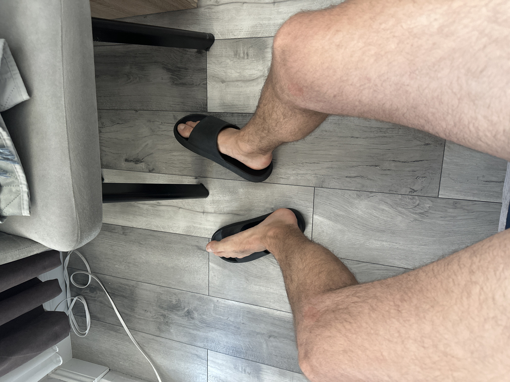 The image shows a person's legs and feet wearing black slides, positioned on a wooden floor. The legs are hairy and tanned. One foot is angled inwards, while the other is pointed more outwards. Behind the legs, a grey upholstered chair is visible, with its legs extending down to the floor. A white cord is coiled on the floor near the chair and curtains. The scene appears to be indoors, possibly in a living space. There is no indication of the specific location within the city of Varna, Bulgaria.