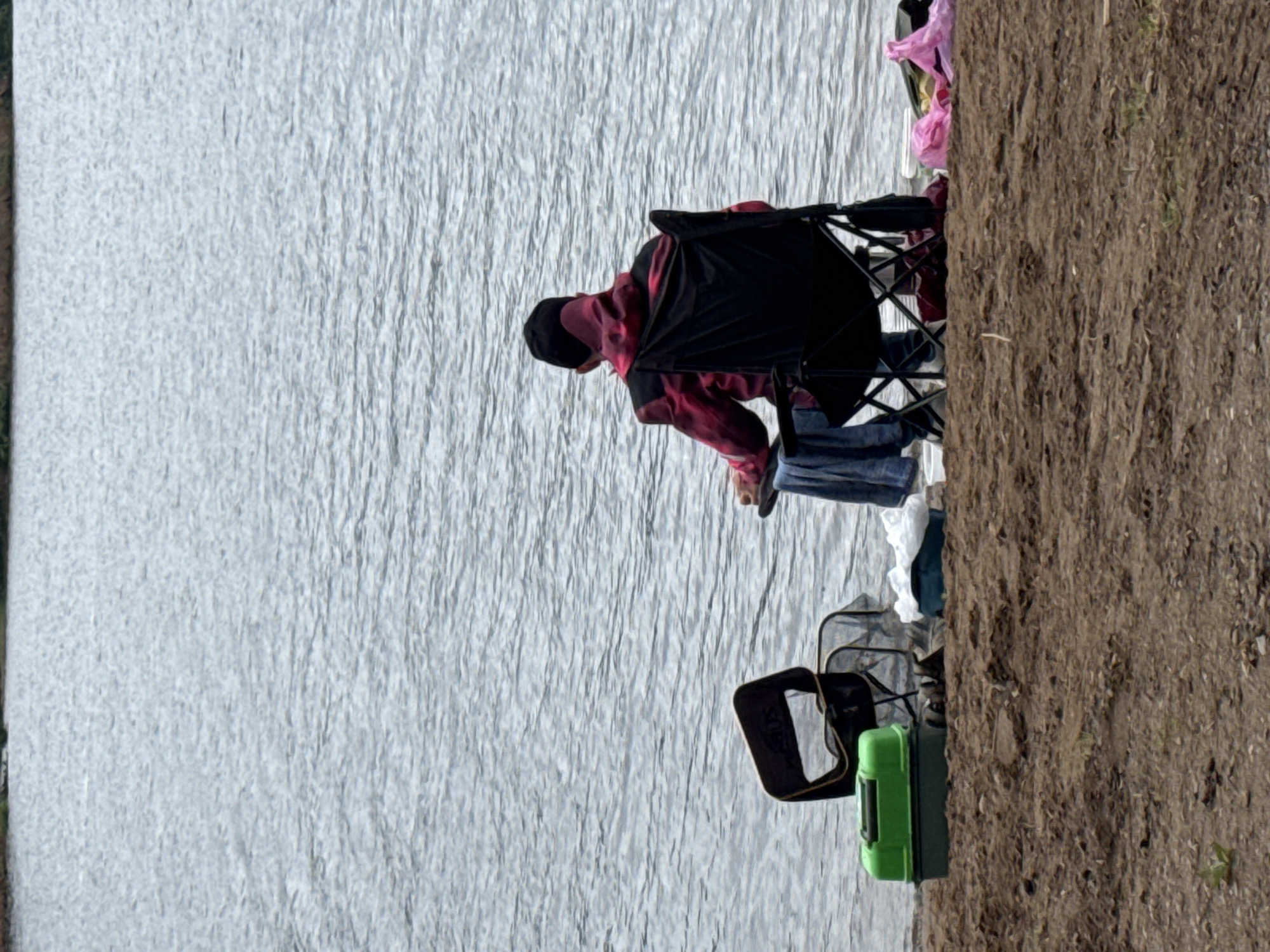 In the city of Chaikovsky, Russia, a single individual is observed from behind, seated on the unpaved bank of a body of water. The subject occupies a black, foldable camping chair. Apparel consists of a black head covering and a long-sleeved garment featuring a black base with irregular red and pink patterns. A blue textile, possibly a towel, is draped over the chair's left armrest.
To the subject's left, on the ground, a green and dark green rectangular fishing tackle box is present. Adjacent to this box, a fishing net, likely a landing net, is partially visible. Further left, a partially folded or empty black chair-like structure is discernible. To the right of the subject's chair, a light-colored container or bucket with white contents is on the ground. A pink plastic bag and additional dark items are also located on the ground further to the right.
The immediate foreground consists of reddish-brown earth, appearing to be a mix of soil, sand, and small pebbles, with minimal sparse green vegetation. The middle and background are dominated by a wide body of water, exhibiting numerous minor surface ripples. A narrow, dark green band along the distant horizon indicates a forested shoreline or landmass. The overall lighting suggests an overcast sky or ambient conditions without direct sunlight. No other individuals or active interactions are depicted.