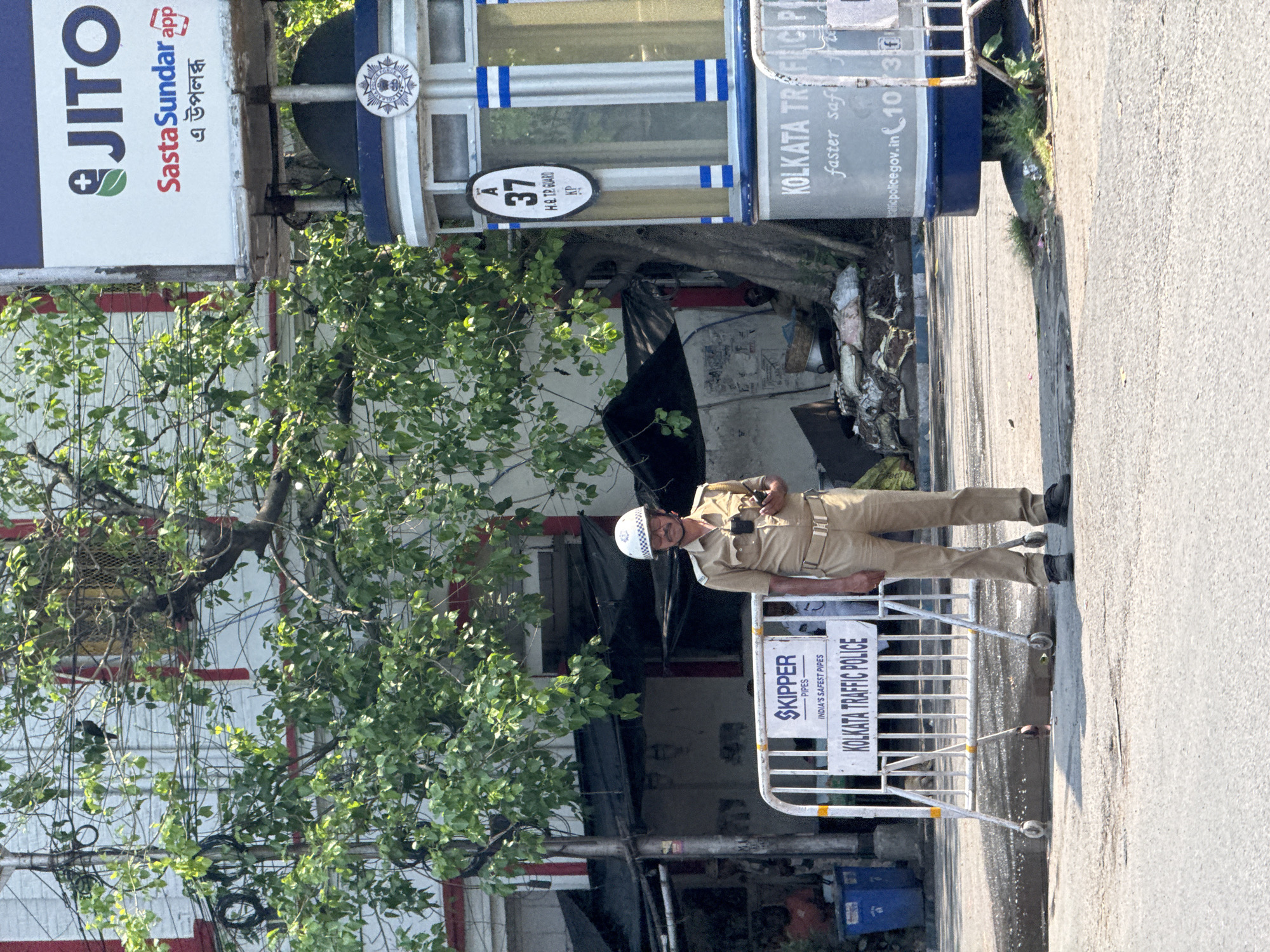 A medium shot captures a Kolkata Traffic Police officer, clad in a khaki uniform and a white helmet, standing behind a white metal barricade. The barricade bears the inscription "SKIPPER PIPES - INDIA'S SAFEST PIPES" and "KOLKATA TRAFFIC POLICE."
Behind the officer and partially obscured by foliage is a Kolkata Traffic Police booth, displaying the number "37" and the text "H.Q. TP. GUARD KP." The booth also features the Kolkata Traffic Police logo and the slogan "faster safe," along with the website "trafficpolice.gov.in" and the phone number "1073."
To the left, a billboard advertises "JITO Sasta Sundar app." The environment suggests a typical urban street scene in Kolkata, India.