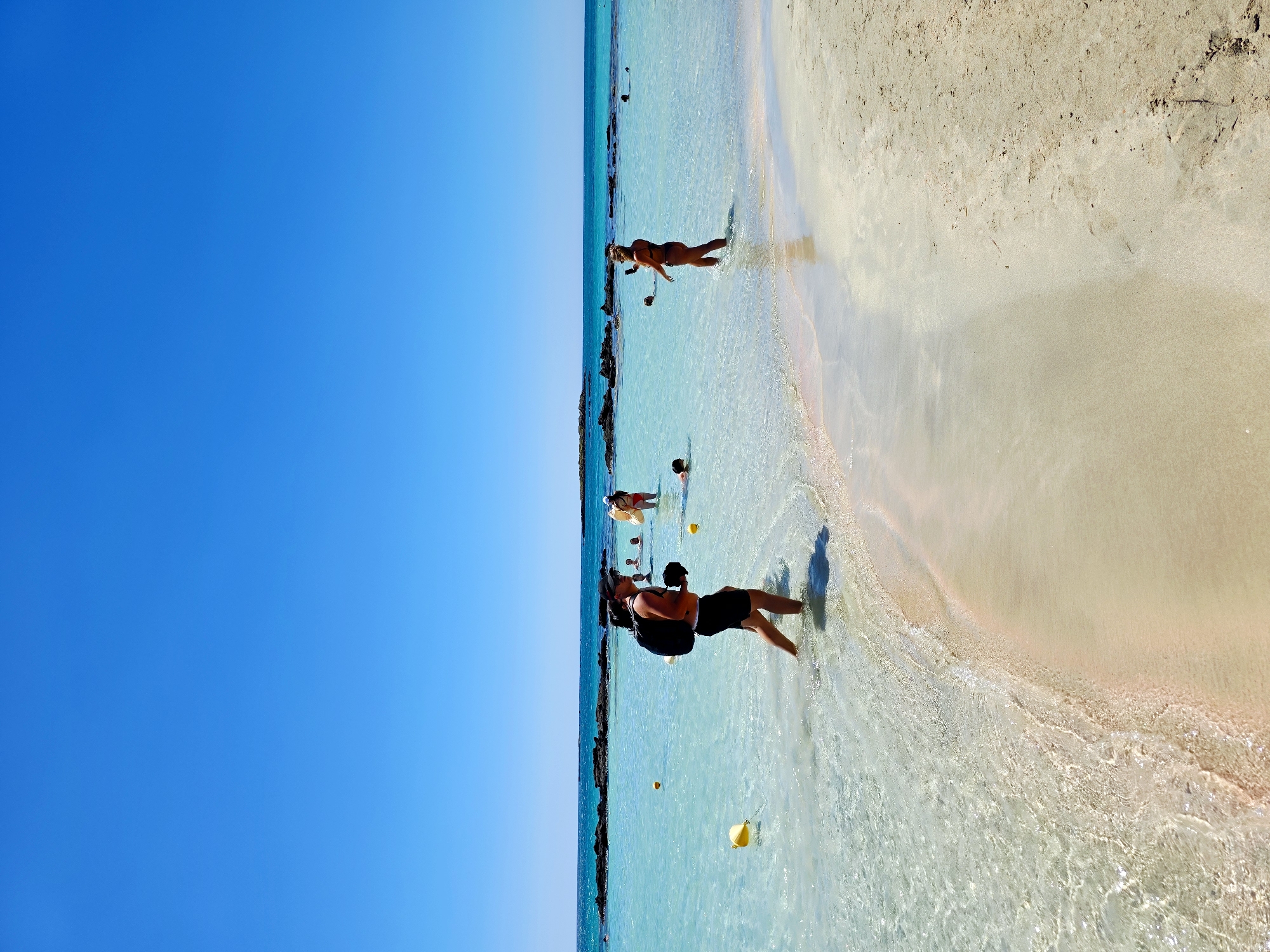 In Elafonissi Beach, Greece, a man with a backpack walks through shallow, turquoise water under a clear blue sky. He is wearing black shorts and a dark cap. Several other people are visible in the water, including a woman in a bikini standing and holding a phone, and another person floating with an inflatable. Yellow buoys are scattered in the water. The sandy beach has a pinkish hue in some areas, and the water is crystal clear, revealing the sandy bottom. Rocky formations are visible in the distance.