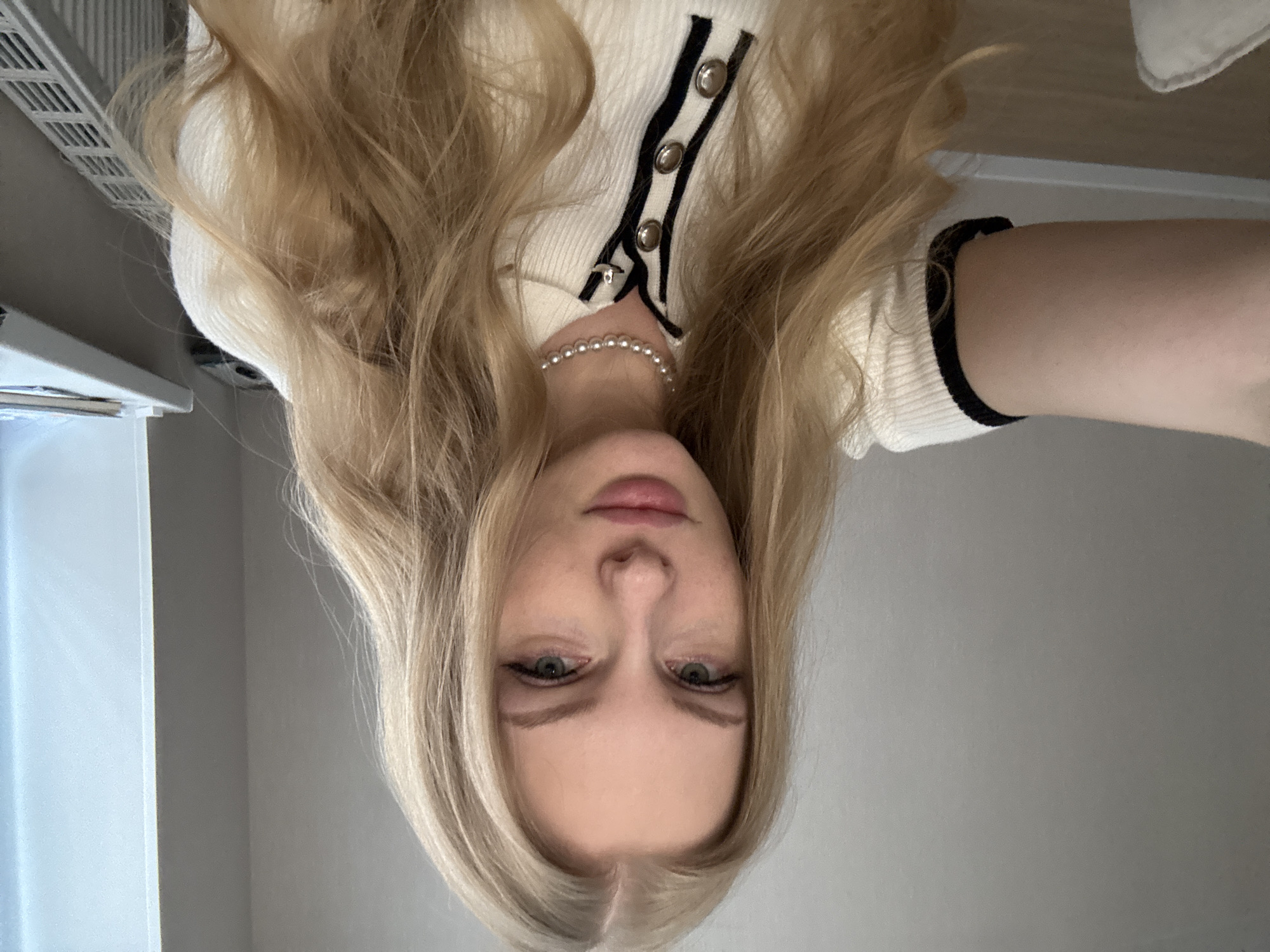A Caucasian female is depicted in a front-facing photograph. She has long, wavy blonde hair with a center part and light-colored eyes. She is wearing a ribbed, cream-colored short-sleeved top featuring a black-trimmed collar and placket with three silver buttons, along with a single-strand pearl choker. Her left arm, consistent with holding a camera for a self-portrait, is visible on the lower-left. The background on the left consists of an unadorned light gray wall. On the right, a window provides natural light, illuminating a windowsill that holds a stack of papers or books and a light-colored electronic device. Below the windowsill, a white panel radiator is visible. The image does not contain any visual information to confirm the location as Perm, Russia.