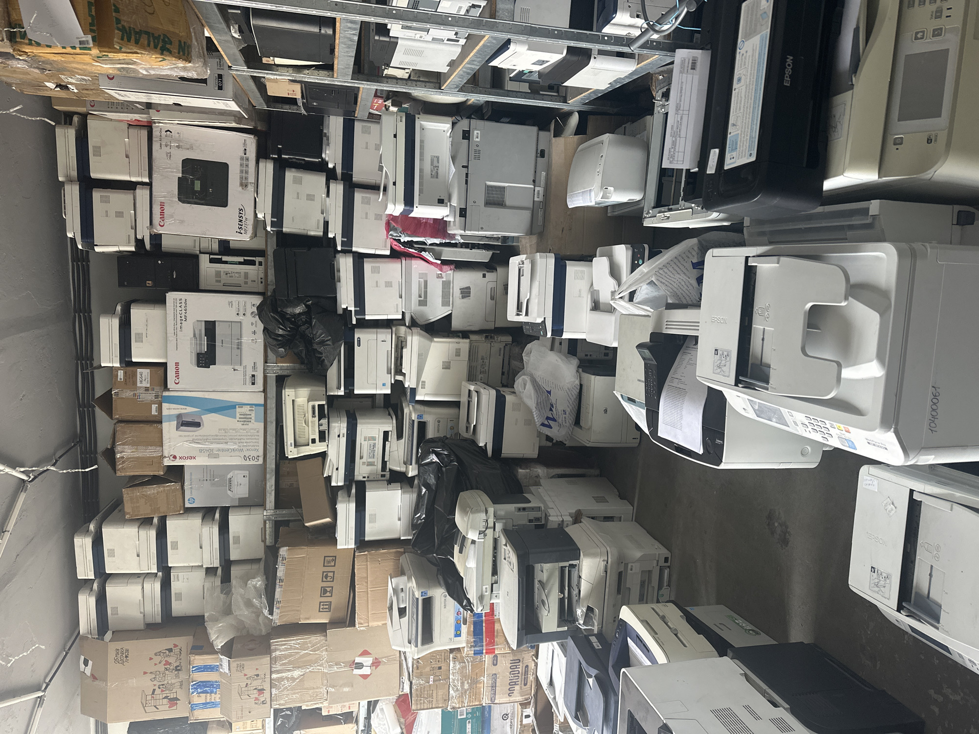 The image depicts a cluttered storage room, presumably in Unknown, filled with numerous printers, both boxed and unboxed. The printers vary in make and model, including brands like Xerox, Canon, and Epson. The objects are stacked haphazardly, some on shelving and others directly on the floor, creating a sense of disorganization and overcrowding. The environment is enclosed, with visible structural elements like walls and shelving supports. The general impression is that of a warehouse or storage facility used for surplus or discarded electronic equipment.