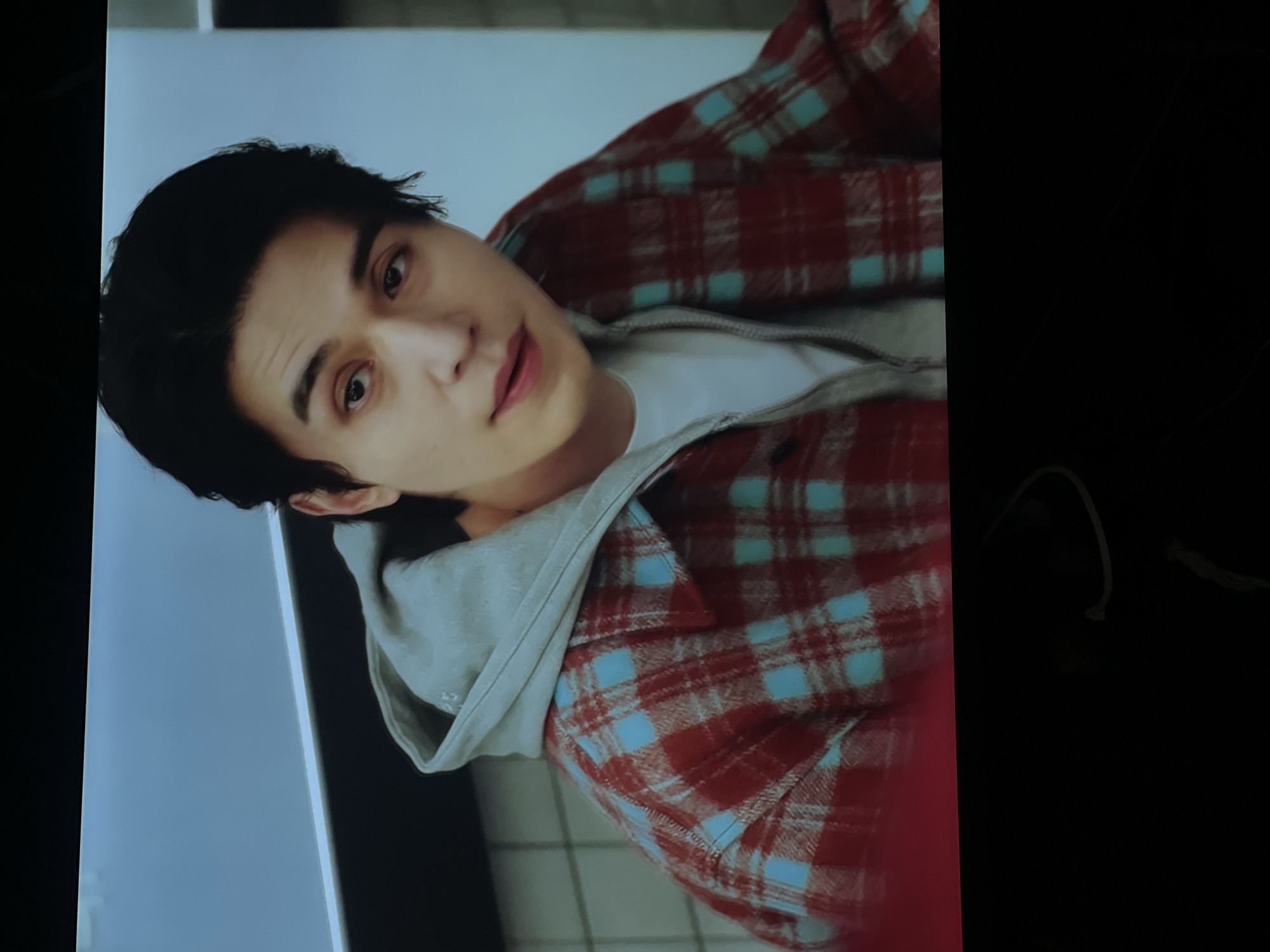A close-up, slightly rotated image shows a man wearing a red and blue plaid shirt over a gray hoodie and a white t-shirt. He has dark hair and a fair complexion. The background is a mix of light blue and black, possibly a wall or a screen. The image appears to be a still from a video or a photograph taken of a screen. There is no indication of the location being in Vologda, Russia.