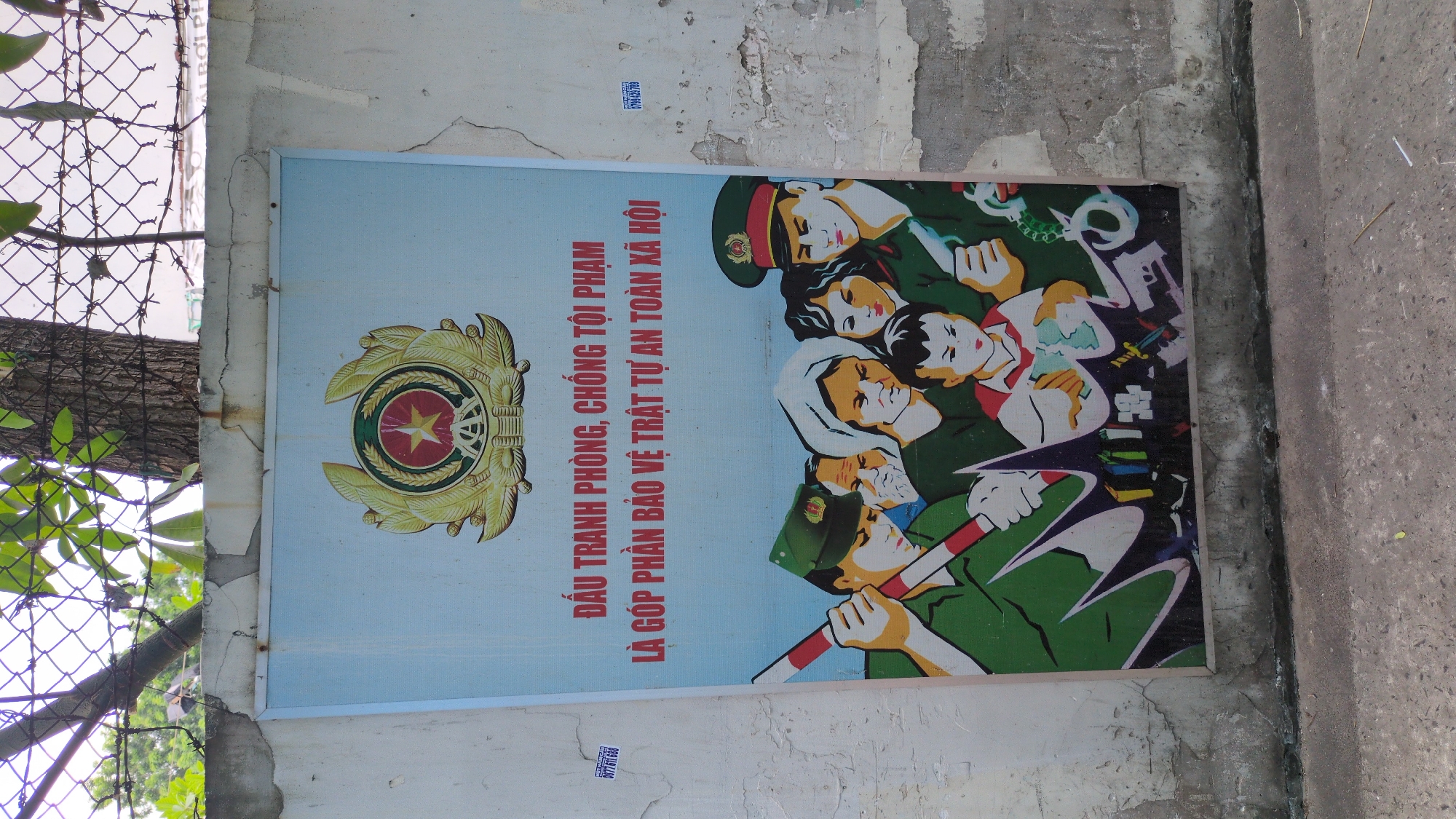 A slightly tilted, medium shot shows a propaganda poster attached to a concrete wall next to a chain link fence with a tree and foliage behind it. The poster has a light blue background with a large, golden emblem in the upper left corner and dark blue text that reads "ĐẤU TRANH PHÒNG, CHỐNG TỘI PHẠM
LÀ GÓP PHẦN BẢO VỆ TRẬT TỰ AN TOÀN XÃ HỘI" to the right of the emblem. The right half of the poster depicts a group of people, including several police officers, in an illustrative style. Some of the officers are holding weapons, and handcuffs are visible in the upper right corner. The surrounding wall shows signs of wear and tear, with cracks and patches of missing paint. Small, rectangular stickers are visible on the wall both above and below the poster, with some text and numbers. The edge of a concrete sidewalk or road is visible in the bottom right corner of the frame. Based on the phone numbers and text on the poster, the image appears to be from the city of Thủ Đức, Vietnam.