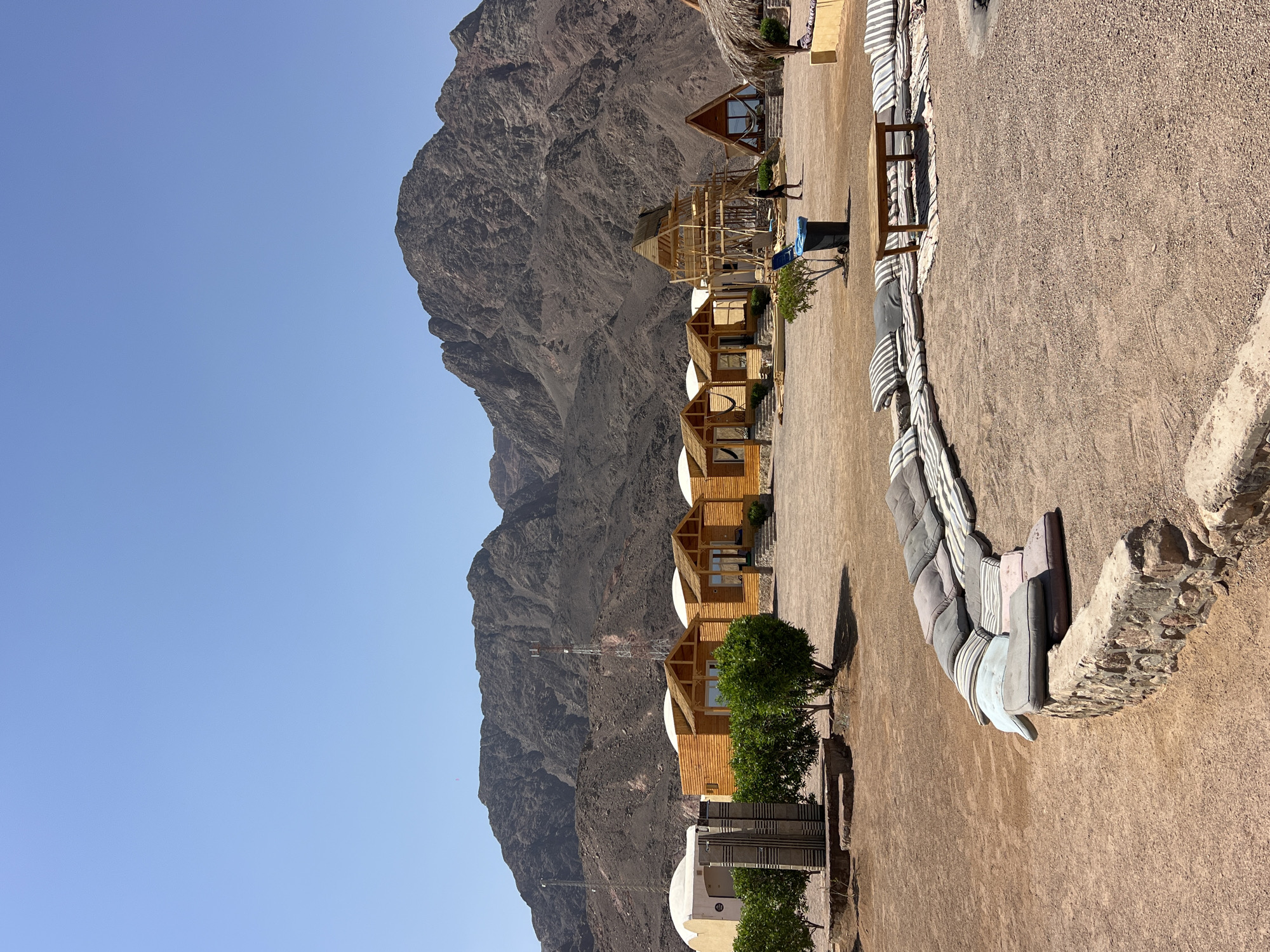 The image depicts a desert resort in Nuweiba, Egypt, with a cluster of wooden bungalows and dome-shaped accommodations set against a backdrop of imposing, arid mountains under a clear blue sky. In the foreground, a low stone wall is topped with an arrangement of floor cushions, forming a seating area around a wooden coffee table placed on a striped rug. Further into the scene, several identical wooden bungalows with thatched roofs are visible, each with a small porch and steps leading to the entrance. One bungalow has a hammock strung across its porch. To the left, a white dome structure is partially visible, adjacent to a tall, abstract sculpture. Further to the right, a wooden structure resembling scaffolding surrounds a partially built triangular hut with a thatched roof. A single person, dressed in dark clothing, walks in the middle ground near a blue trash bin. In the distance, a telecommunications tower stands on the mountainside. The ground is a mixture of sand and small stones, characteristic of a desert environment. The overall impression is one of a serene, rustic retreat in a remote desert landscape.