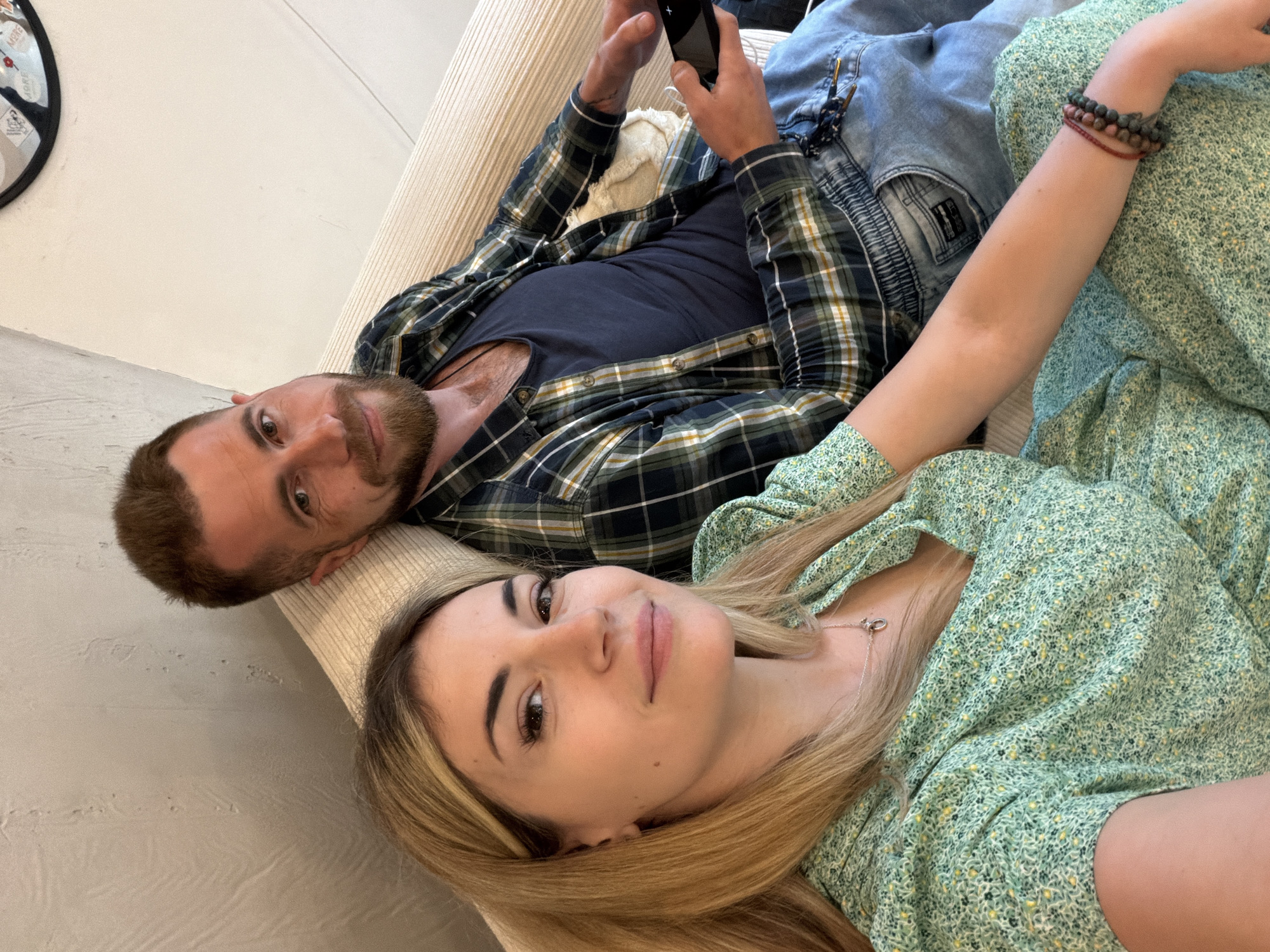 Two individuals, a man and a woman, are lying down indoors, likely on a couch. The man is positioned towards the left side of the frame and is holding a phone, while the woman lies beside him, facing upward. The man is wearing a navy blue t-shirt under a plaid shirt, and jeans. The woman is wearing a green floral-patterned dress and a bracelet. The environment appears to be inside a residence, though the specific location in Budva, Montenegro, cannot be determined without additional context.