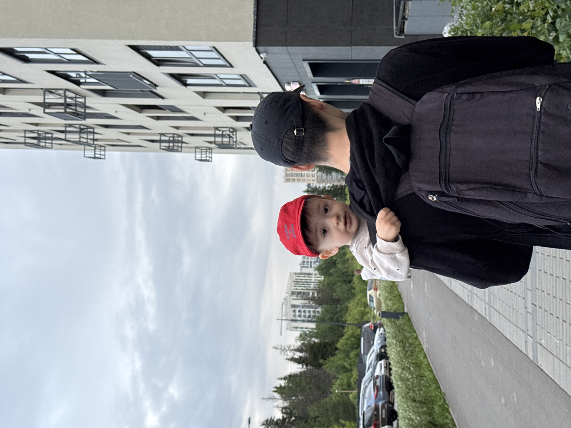 An adult, viewed from behind, wearing a black baseball cap, a black hooded sweatshirt, and a black backpack, carries a small child on their left shoulder. The child, wearing a red baseball cap and a light-colored long-sleeve top, looks directly at the camera. They are walking on a paved sidewalk, with a row of parked cars visible to their left, beyond a strip of green bushes. In the background to the left, dense green trees obscure some distant buildings, with another multi-story residential building and a construction crane partially visible. To the right, a modern, light-colored multi-story residential building with numerous windows and small, square, dark metal balcony structures extends upwards. The sky is overcast. This scene is located in Izhevsk, Russia, specifically within the vicinity of the residential complex "Akvareli" (ЖК "Акварели"), which features buildings with this distinctive architectural style, along or near Ulitsa Pushkinskaya 245B.