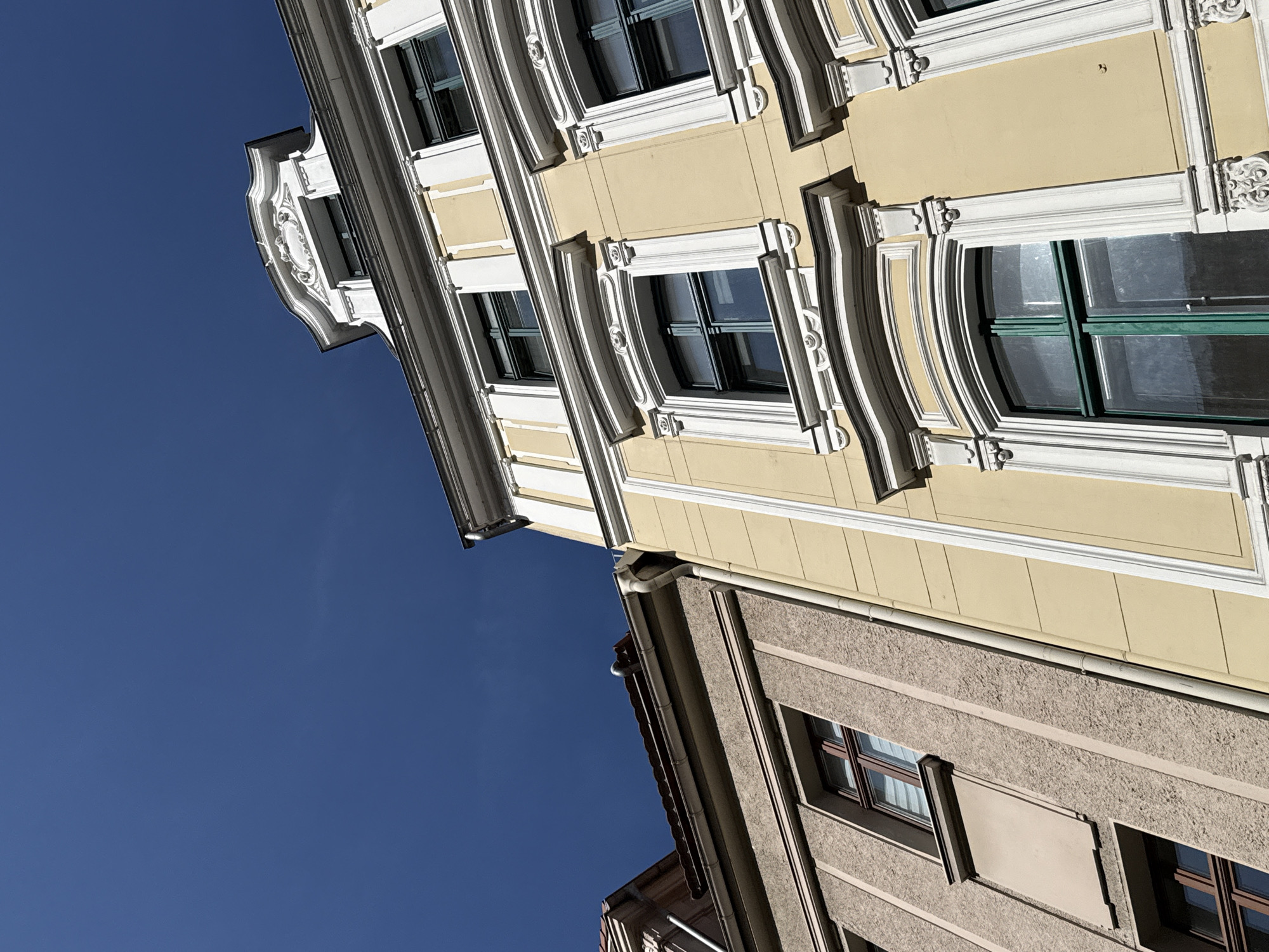 This is a low-angle shot of two buildings in Görlitz, Germany, against a clear blue sky. The building on the right is painted yellow and features white decorative trim around its windows and roofline. It has multiple stories, each with large windows framed by ornate white detailing. The building on the left is a lighter tan color with a rougher texture and has simpler window frames. The tops of both buildings, including roofs and gutters, are visible. The sunlight casts shadows, indicating it is daytime.
